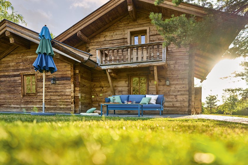 Weishäupl Klassiker Schirm als Sonnenschutz vor einem Holzhaus mit blauer Outdoor-Couch und Gartentisch.