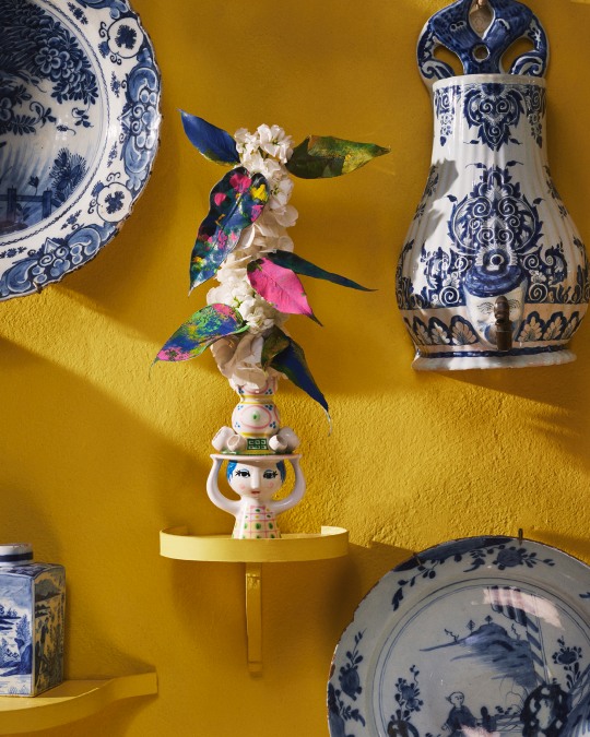 Dekorativer Bjorn Wiinblad Kerzenständer "Lady with Hat" auf gelber Wand mit Blumen und blau-weißen Keramiken.