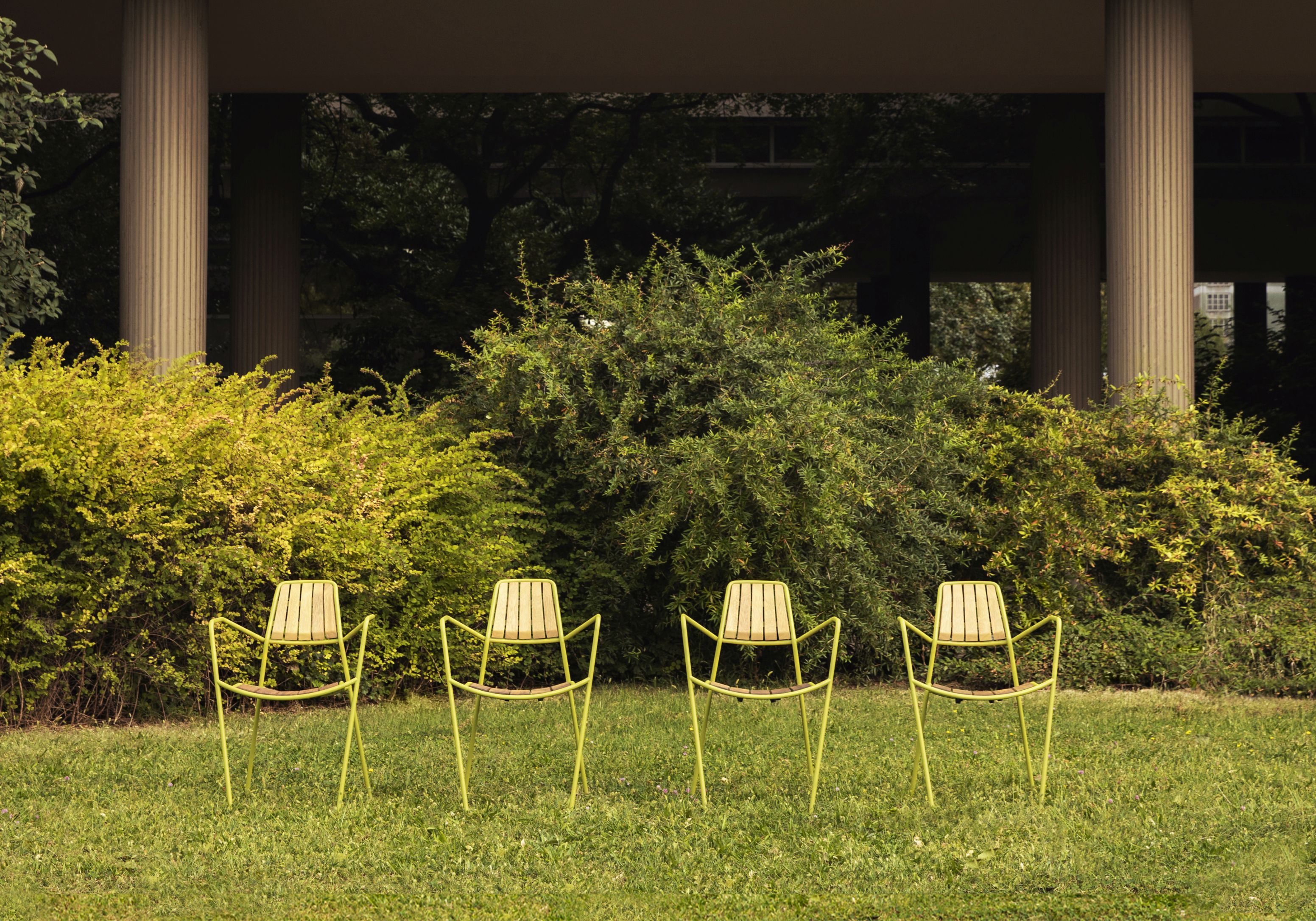 Vier Osmo Outdoor Stühle von Prostoria in Grün auf Rasen vor Büschen und Säulen.
