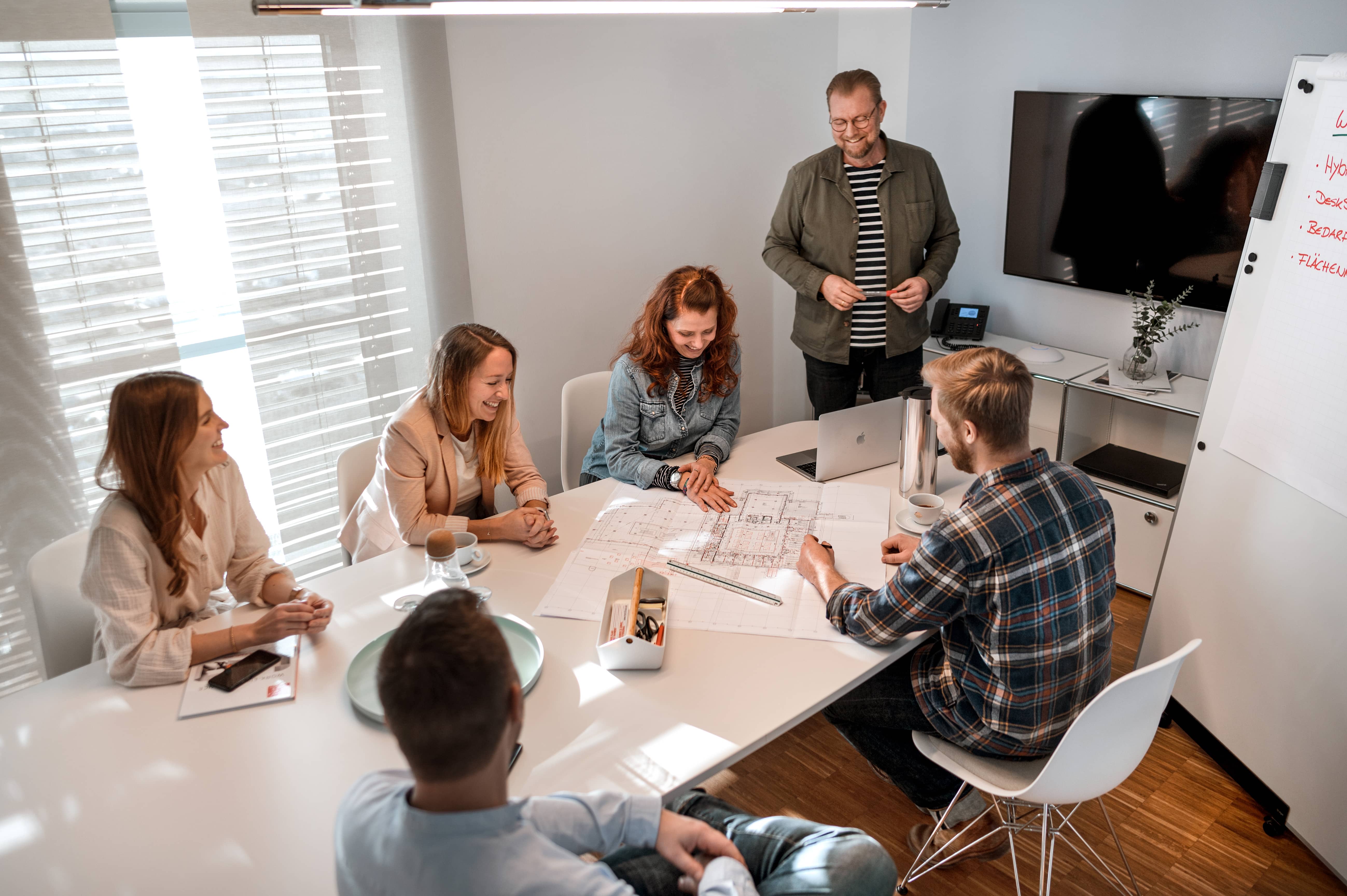 Architekten-Team bespricht Bauplan am Tisch im Büro, mit Laptop, Kaffee und Zeichenutensilien.