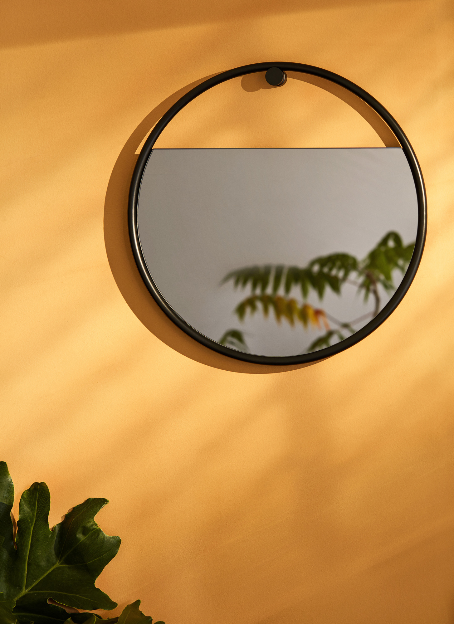 Peek Wandspiegel rund mit schwarzem Rahmen an ockerfarbener Wand, spiegelt Pflanze wider.