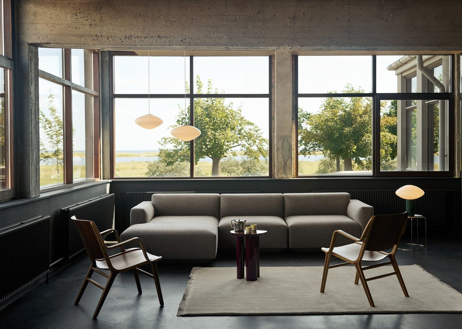 Wohnzimmer mit großem Fenster, grauem Sofa, Holzstühlen und AndTradition The Moor Teppich in Beige.
