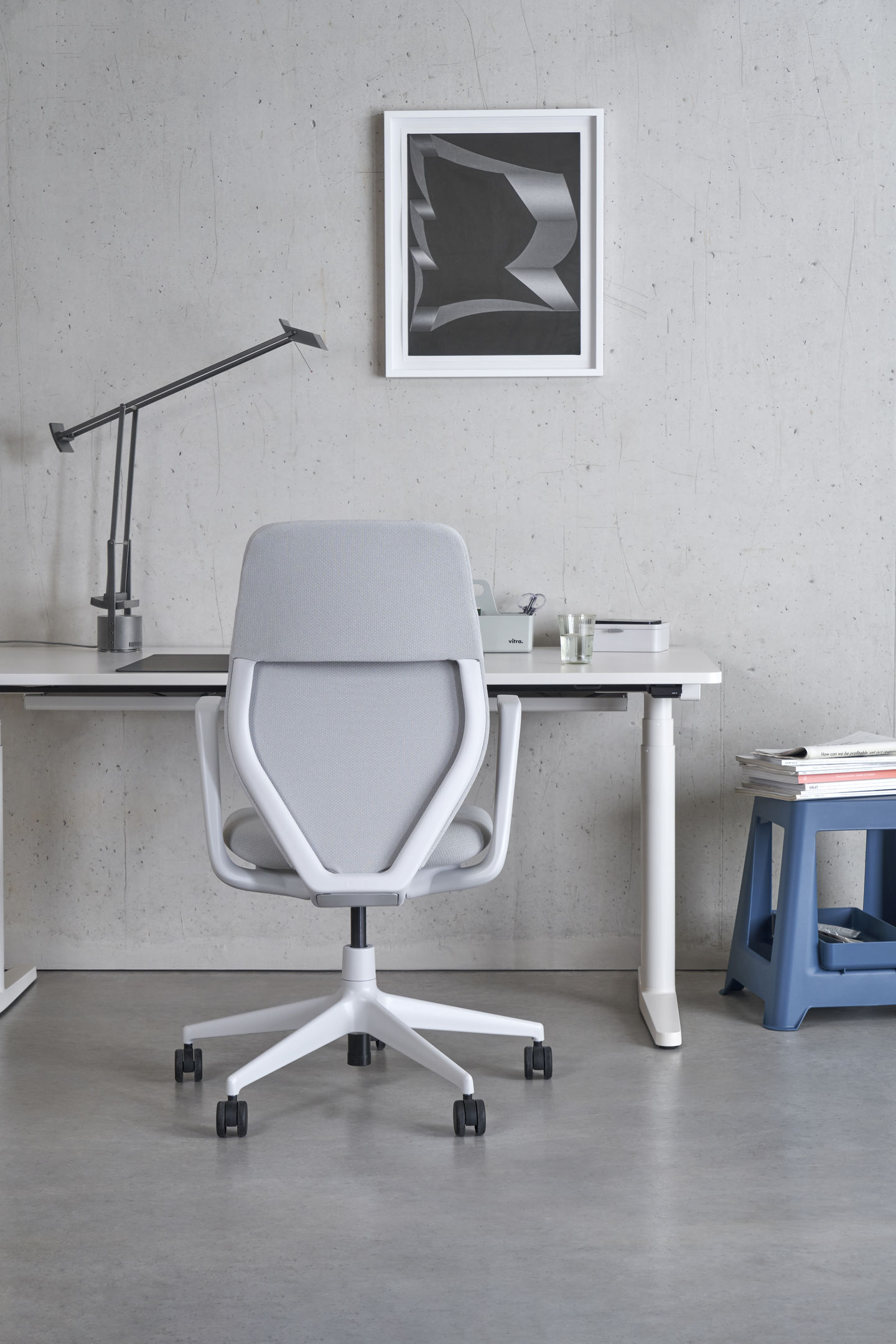 Vitra Tyde 2 Steh-/Sitztisch Home Desk mit Bürostuhl, Lampe und Dekoration vor grauer Wand.