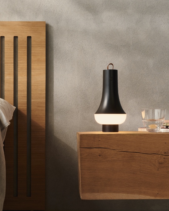 Schwarze Nachttischlampe mit Ledergriff auf einem Holznachttisch neben einem Bett im Schlafzimmer.