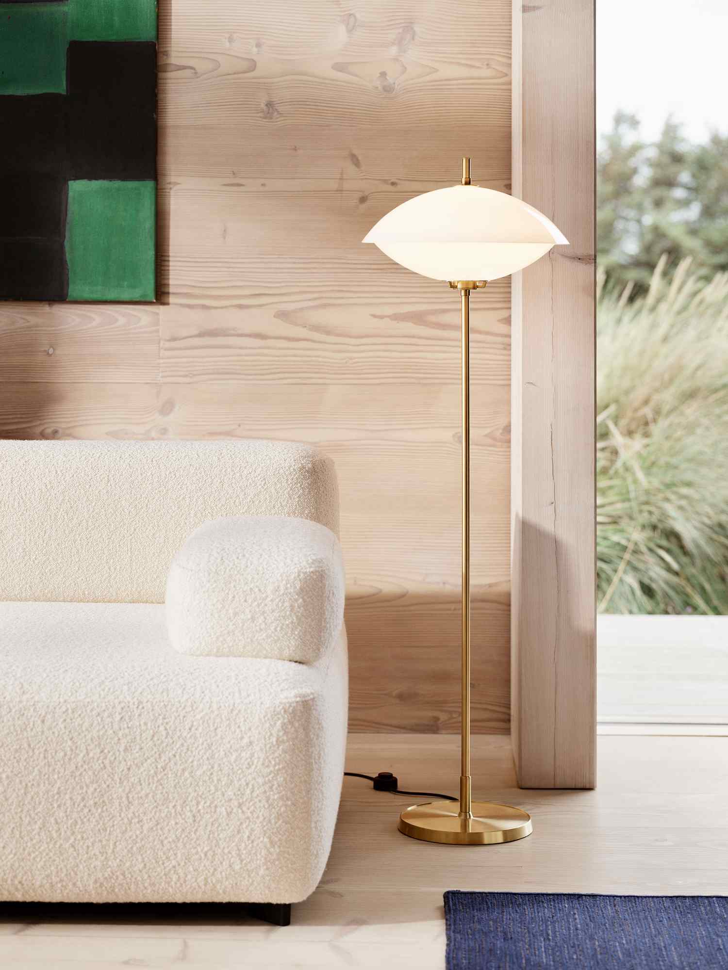 Goldene Stehleuchte von Fritz Hansen mit weißem Schirm neben einem cremefarbenen Sofa.