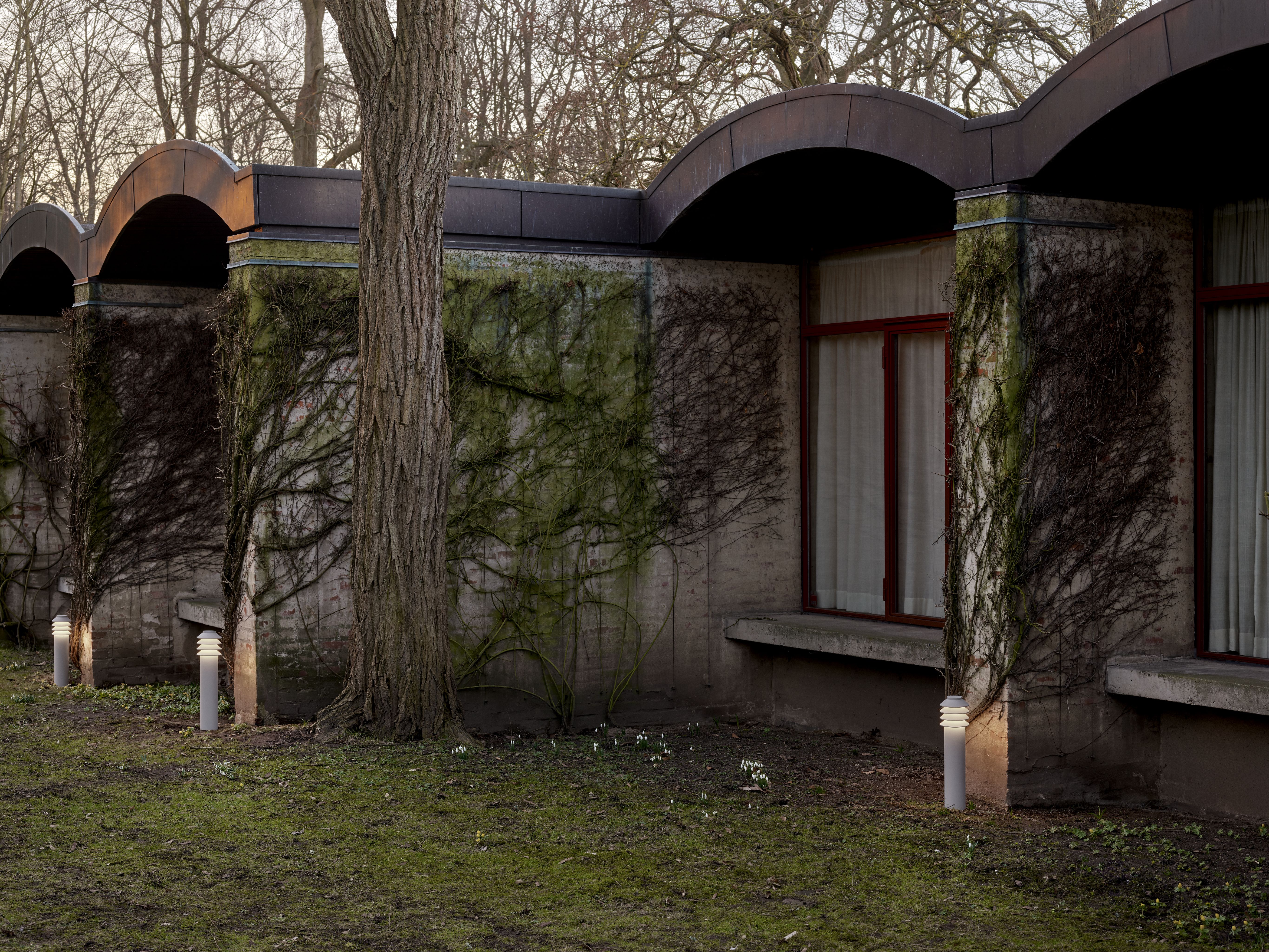 Bysted Garden Pollerleuchte von Louis Poulsen im Außenbereich, stilvolle Gartenbeleuchtung neben einem Gebäude mit Bewuchs.