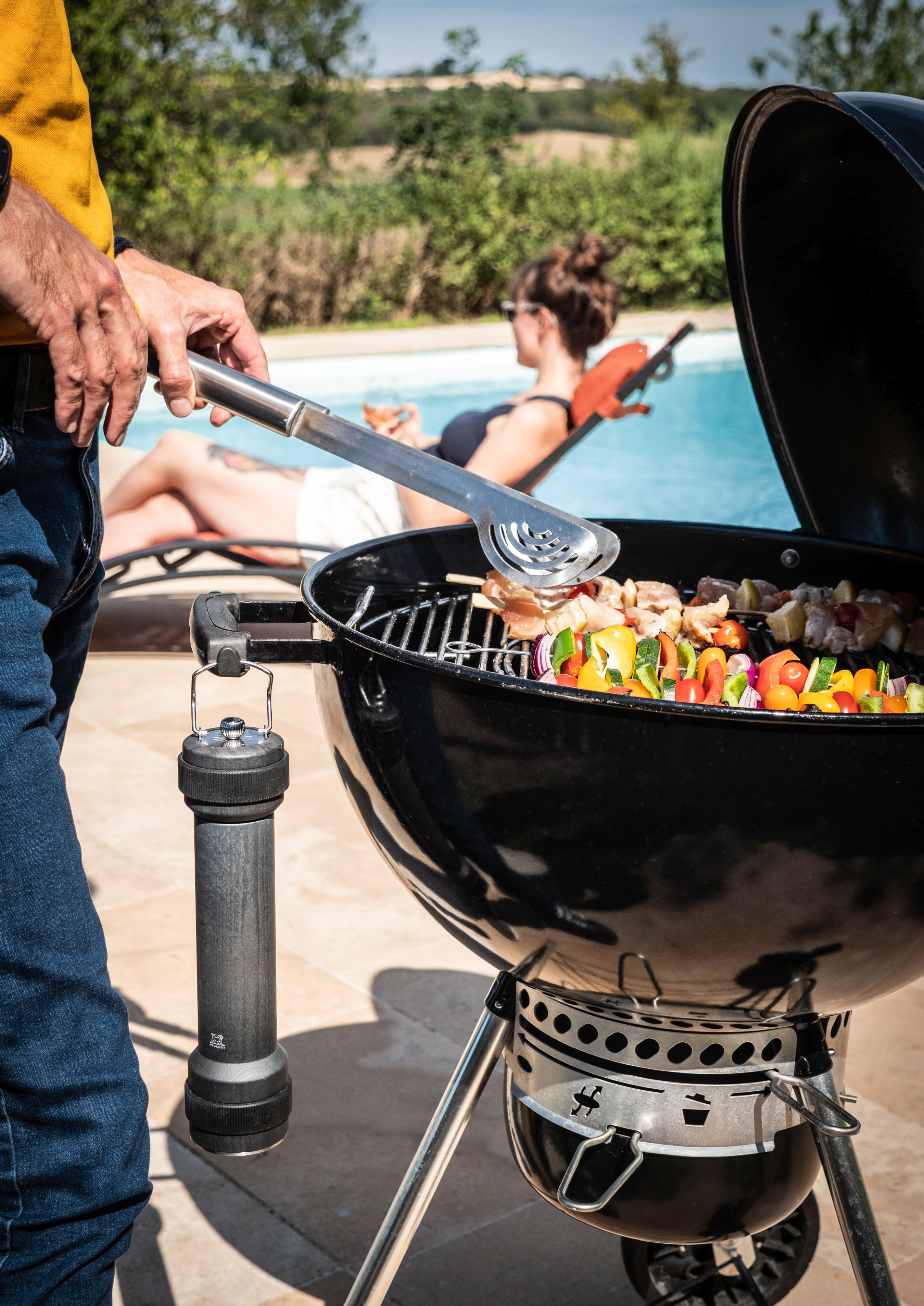 BBQ Szene mit graphitfarbener Peugeot Pfeffermühle, Grillgut auf dem Rost und entspannter Frau am Pool.