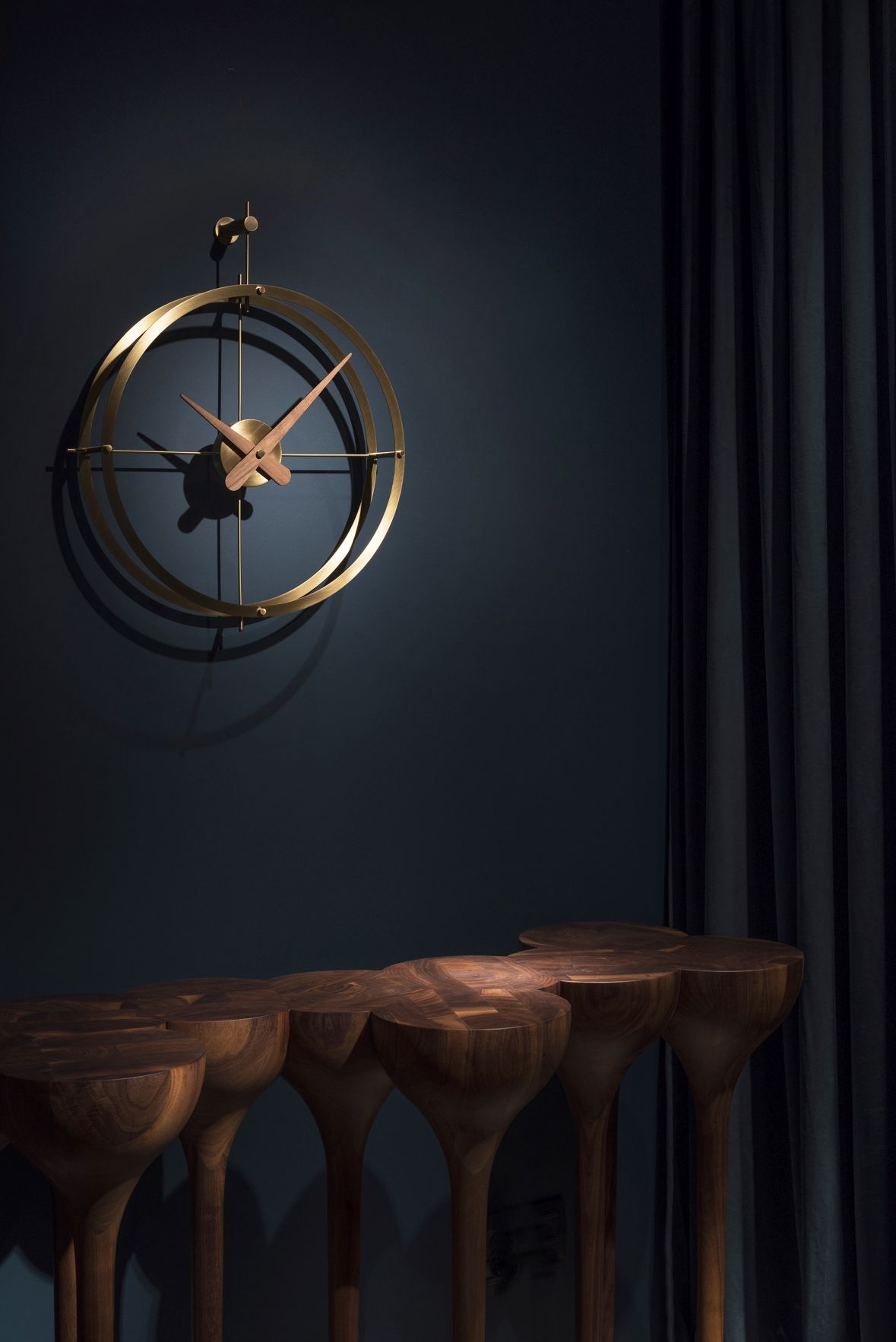 Nomon 2 Puntos Gold Uhr, moderne Wanduhr mit goldenem Rahmen und Holzzeigern vor dunkler Wand.