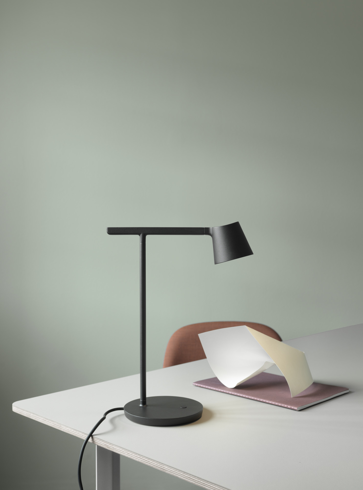 Schwarze Tip Table Lamp Tischleuchte von Muuto auf einem Schreibtisch mit Notizbuch vor hellgrünem Hintergrund.