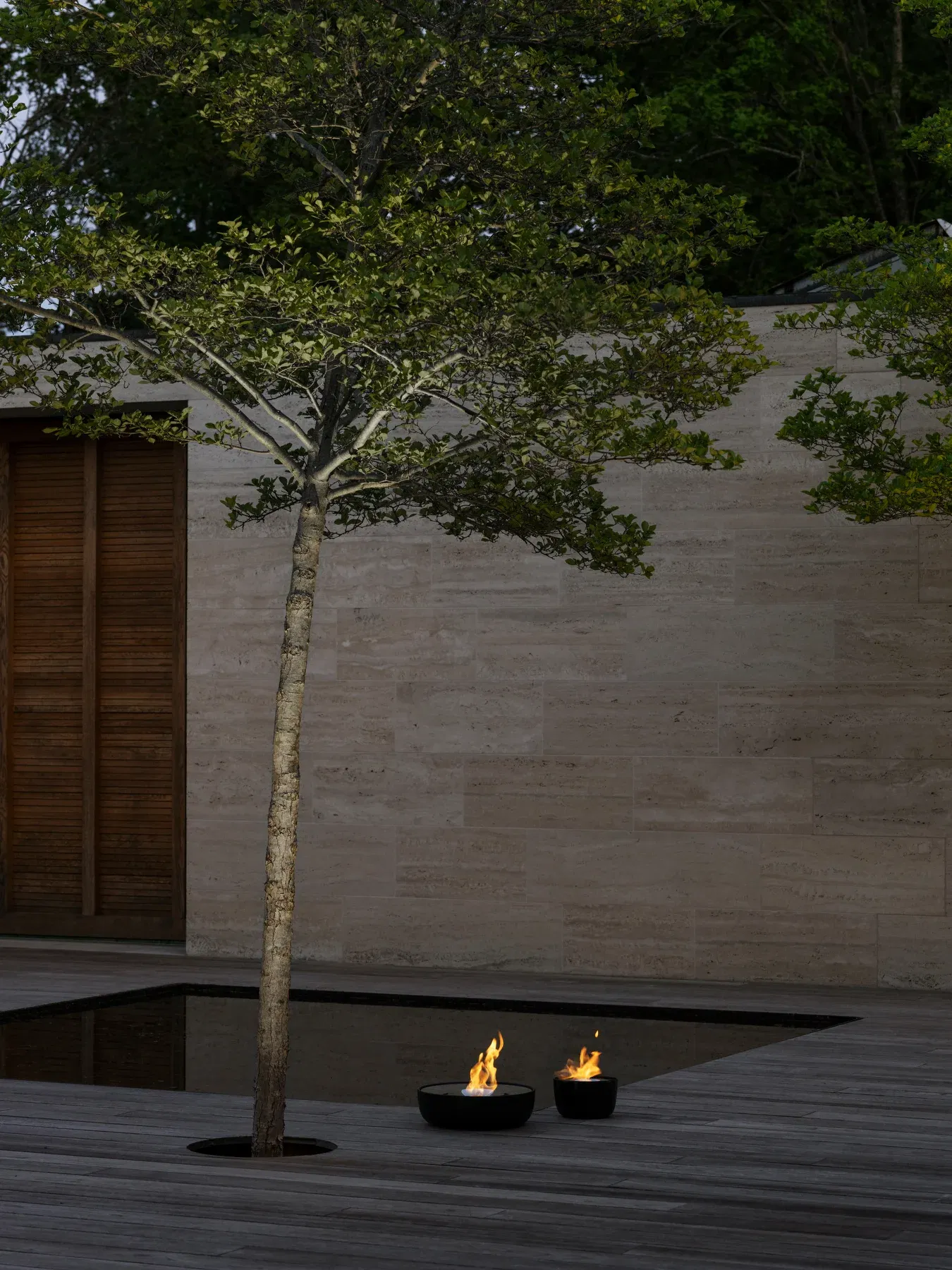 Zwei schwarze Feuerstellen auf Holzterrasse mit Baum und Wasserbecken für stimmungsvolle Gartenbeleuchtung.