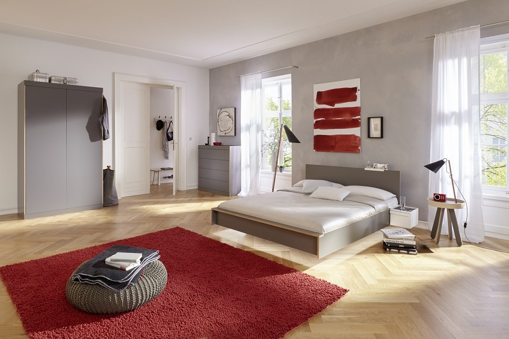 Helles Schlafzimmer mit modernem Flai Kleiderschrank von Müller Möbelwerkstätten, Bett, rotem Teppich und Fenster.
