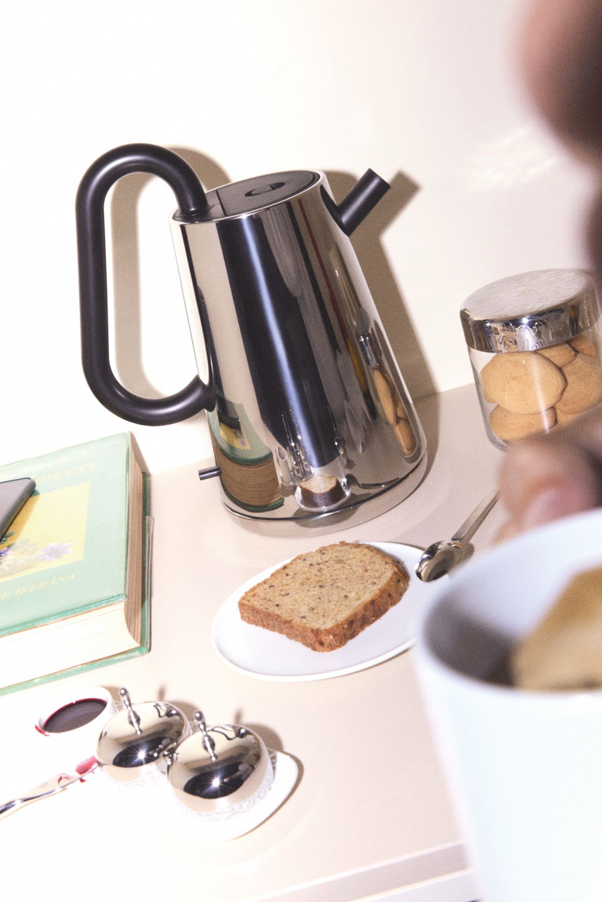 Alessi Toru Wasserkocher 80cl aus Edelstahl auf Tisch mit Tasse, Brot und Keksen.