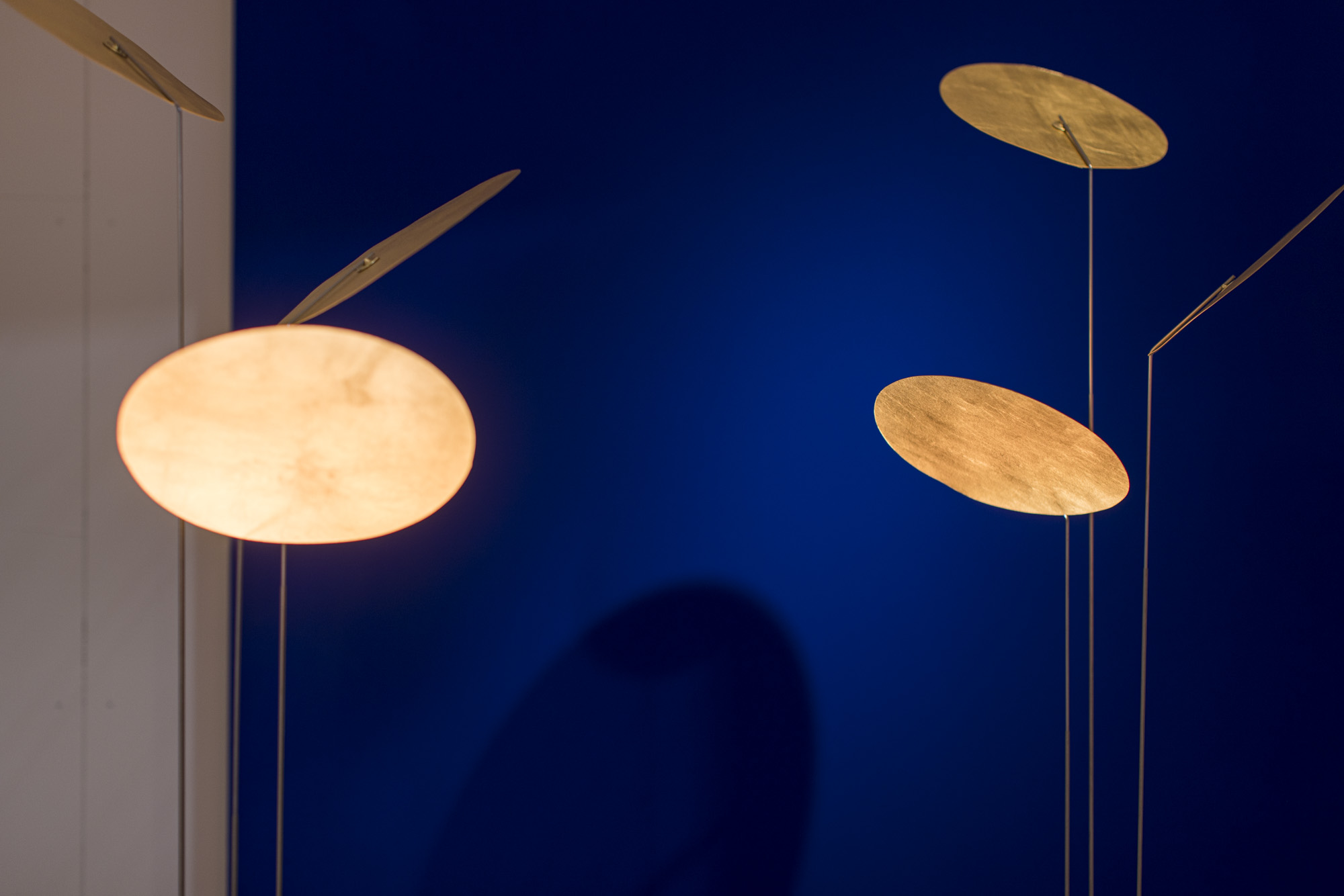 Nahaufnahme der Nuunu Stehleuchte von Ingo Maurer mit goldenen, schwebenden Reflektoren vor blauem Hintergrund.