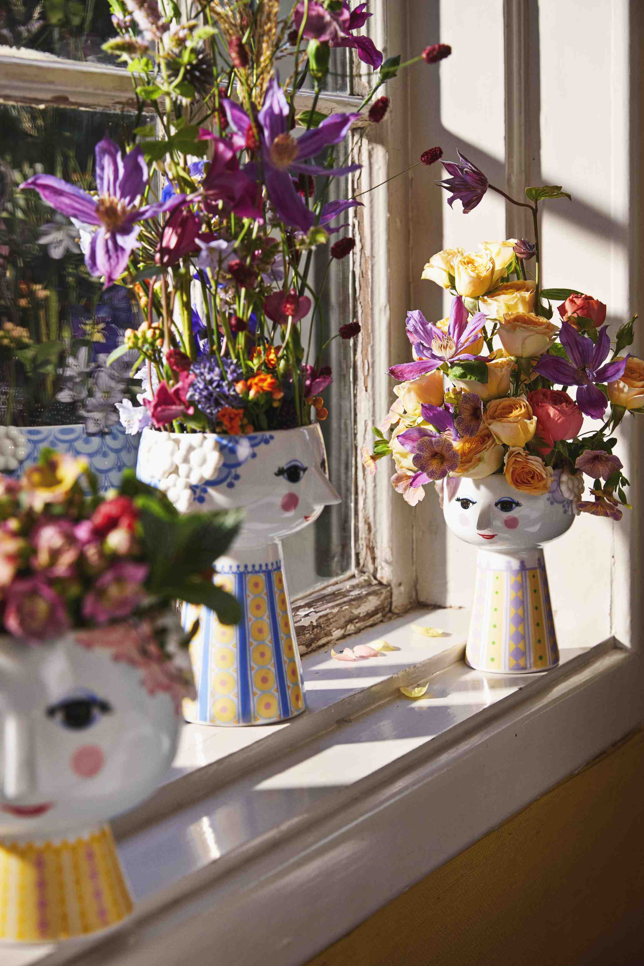 Drei bunte Keramikvasen mit Gesichtern, gefüllt mit frischen Blumen, auf einer Fensterbank. Dekorative Blumenvase.