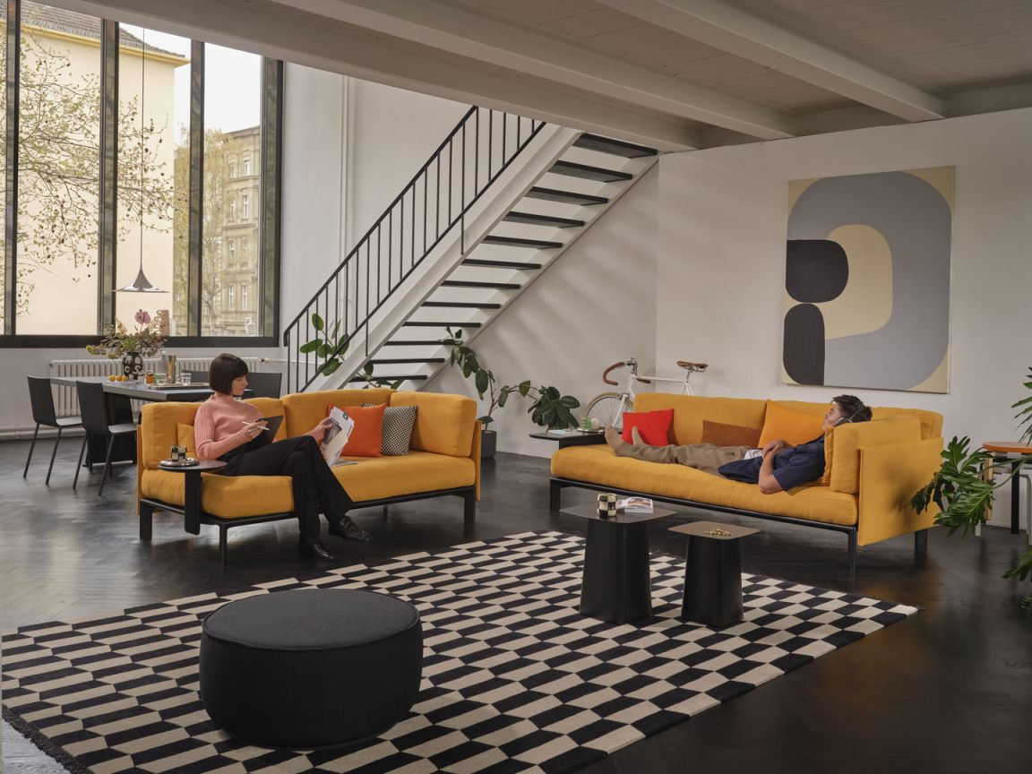 Wohnzimmer mit zwei gelben Sofas mit Anagram Rückenkissen und Personen, die sich entspannen.