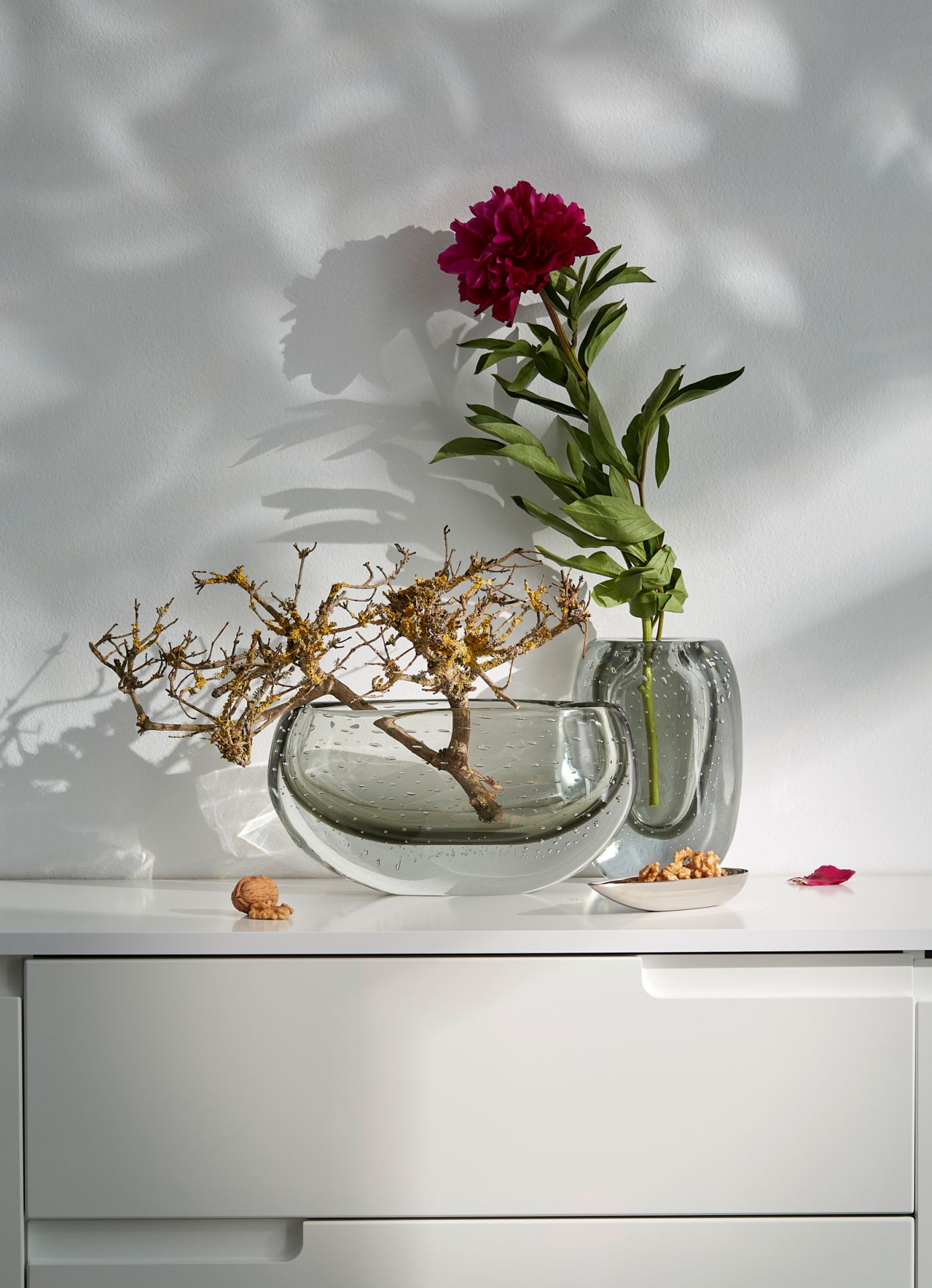 Zwei graue Glasvasen mit Blüte und Zweig als Dekoration auf einer weißen Kommode.