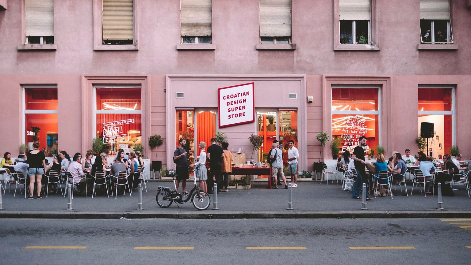 Außenansicht eines Cafés mit Prostoria Stühlen, Tischen und Gästen vor einem rosa Gebäude.
