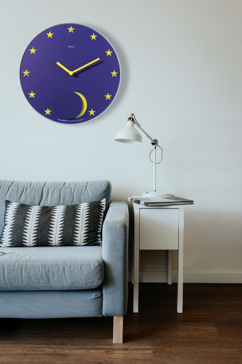 Appuntamento Wanduhr von Rexite in blau mit gelben Akzenten an einer weißen Wand.