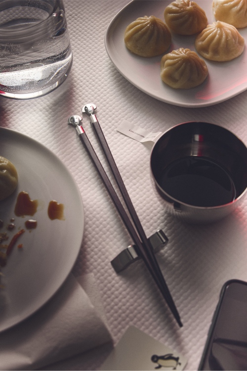Alessi Anna Stick AM44SET1 Essstäbchen auf gedecktem Tisch mit Dim Sum und Sojasauce.