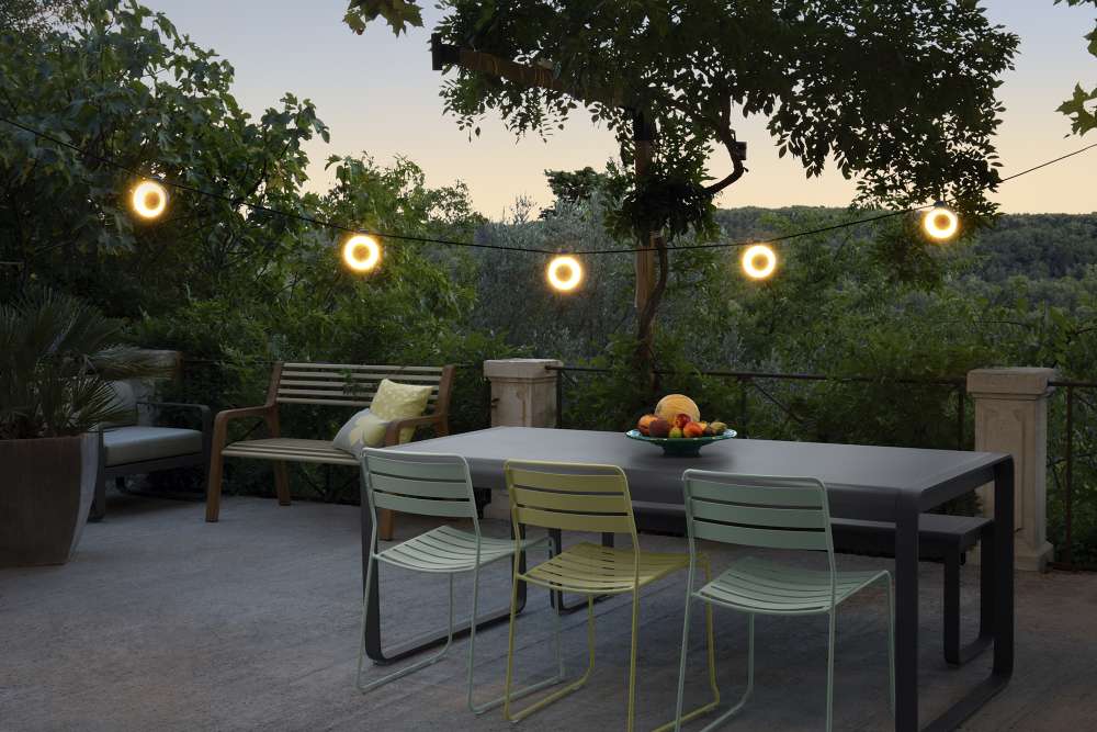 Somerset Bank Outdoor Fermob: Terrasse mit Gartenmöbeln, Tisch, Stühlen, Bank und Lichterkette im Grünen.