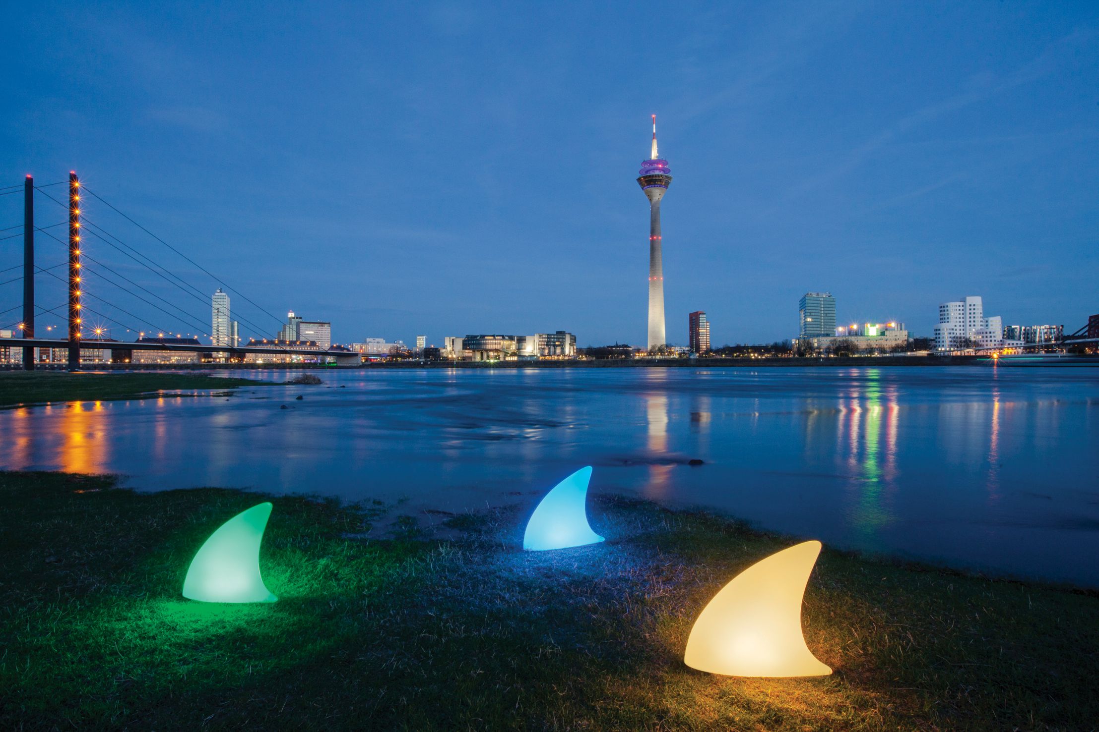 Moree Shark Haifischflosse Bodenleuchte, drei farbige LED-Leuchten am Flussufer mit Skyline im Hintergrund.