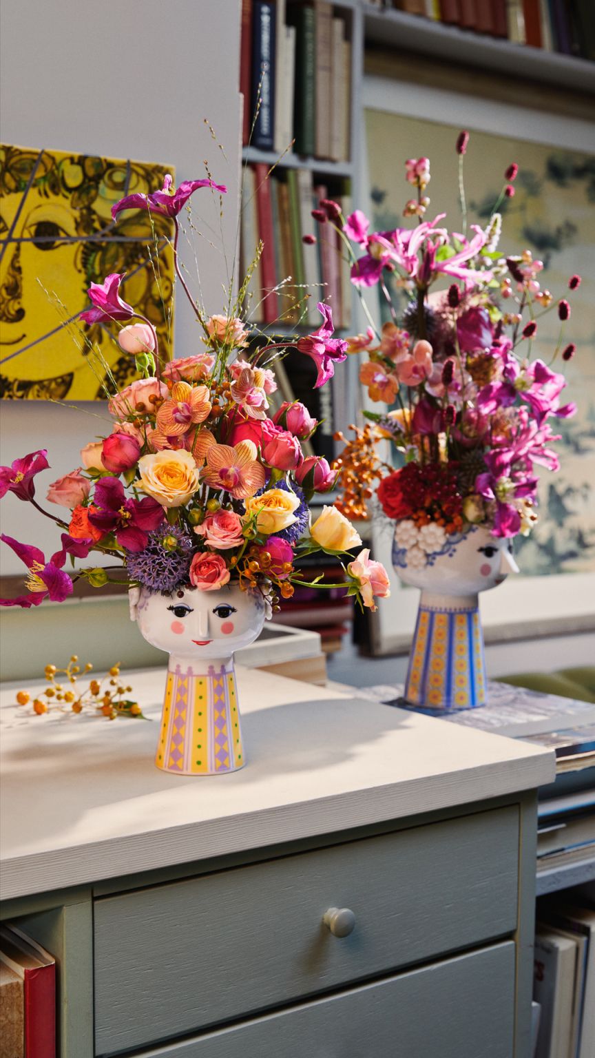 Zwei hellblaue Bjørn Wiinblad Eva Vasen (H18,5cm) mit bunten Blumenarrangements auf einem Tisch.