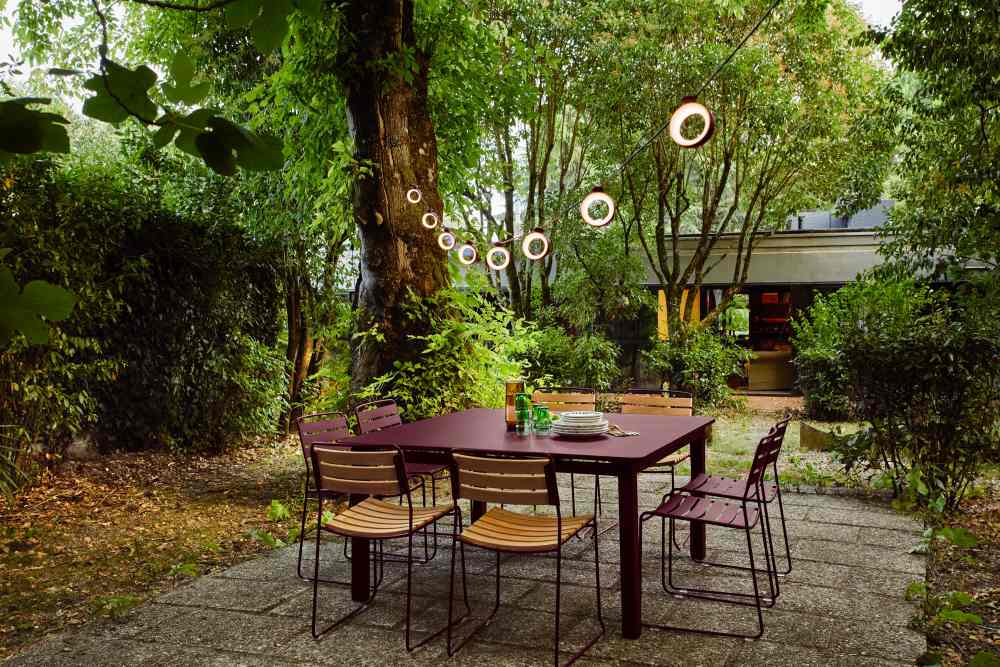 Fermob Surprising Teak Chair Outdoor: Gartenszene mit Teakholz Stühlen und Esstisch unter Lichterketten im Garten.