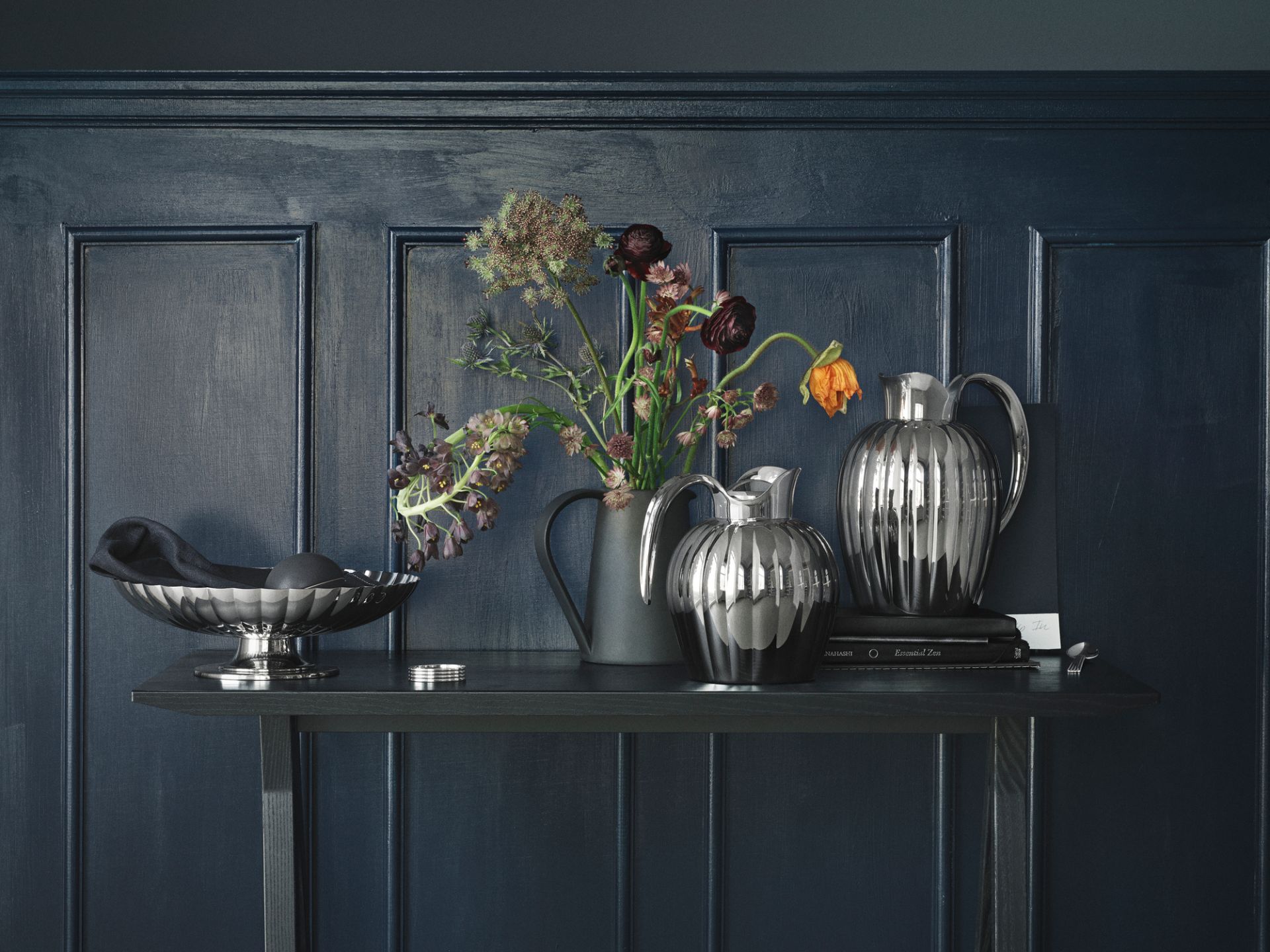 Silberne Karaffen und Schale auf dunklem Regal vor blauer Wand, elegante Tischdekoration.