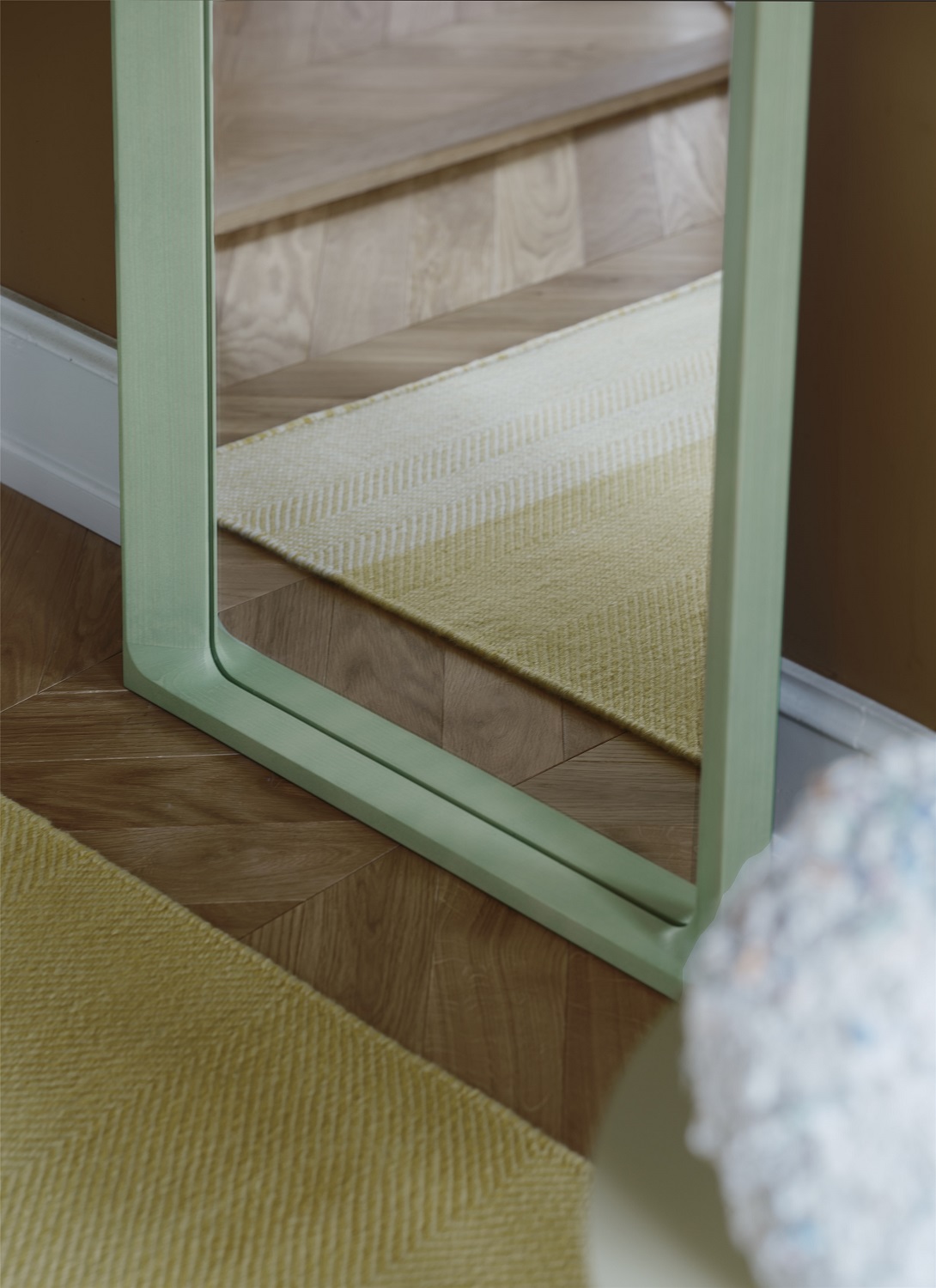 Detailaufnahme des Muuto Arced Mirror Spiegels mit hellgrünem Rahmen, der einen Teppich auf einem Holzboden reflektiert.