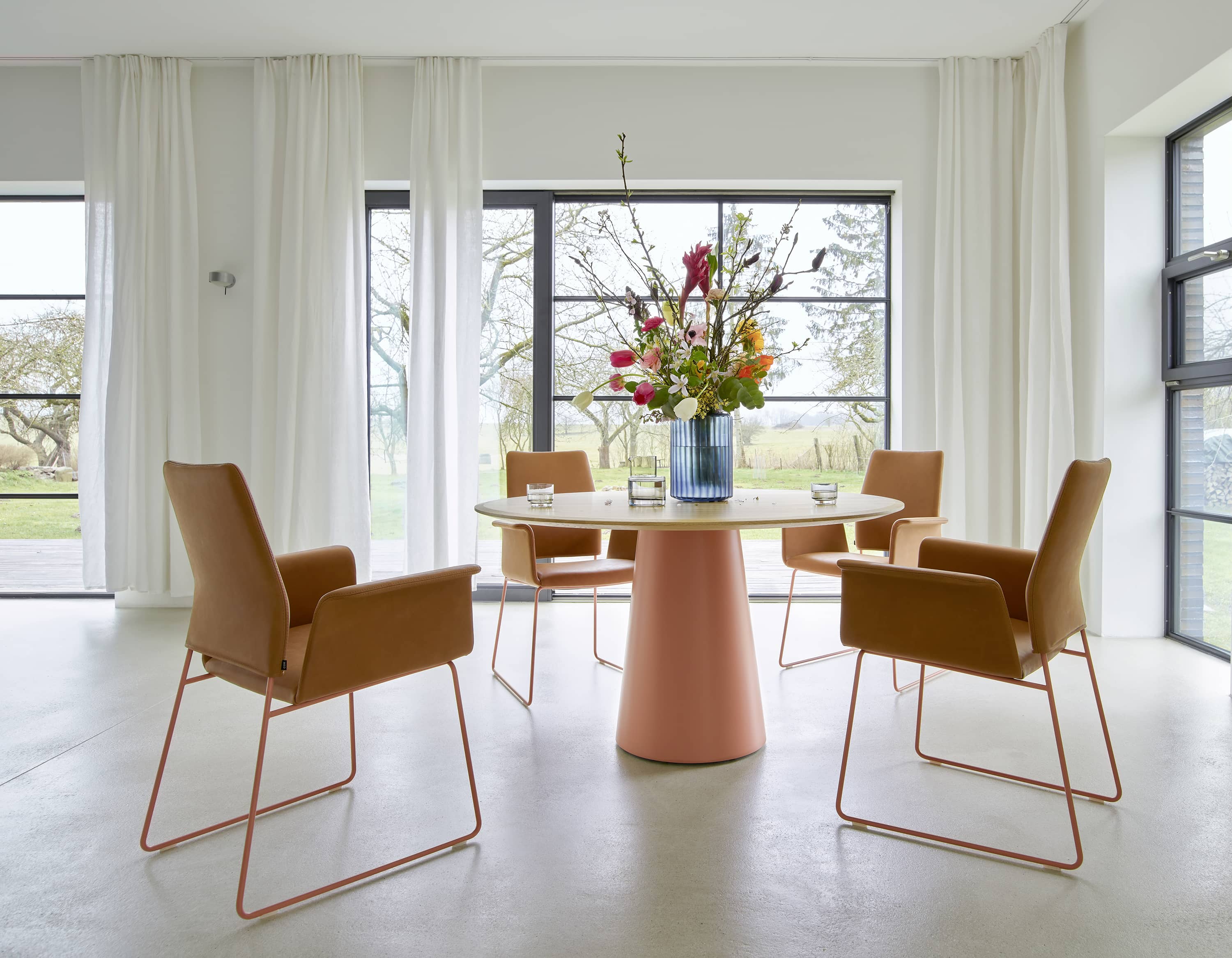 Esszimmer mit rundem Tisch, vier braunen Stühlen und Blumen in blauer Vase vor Fenster.