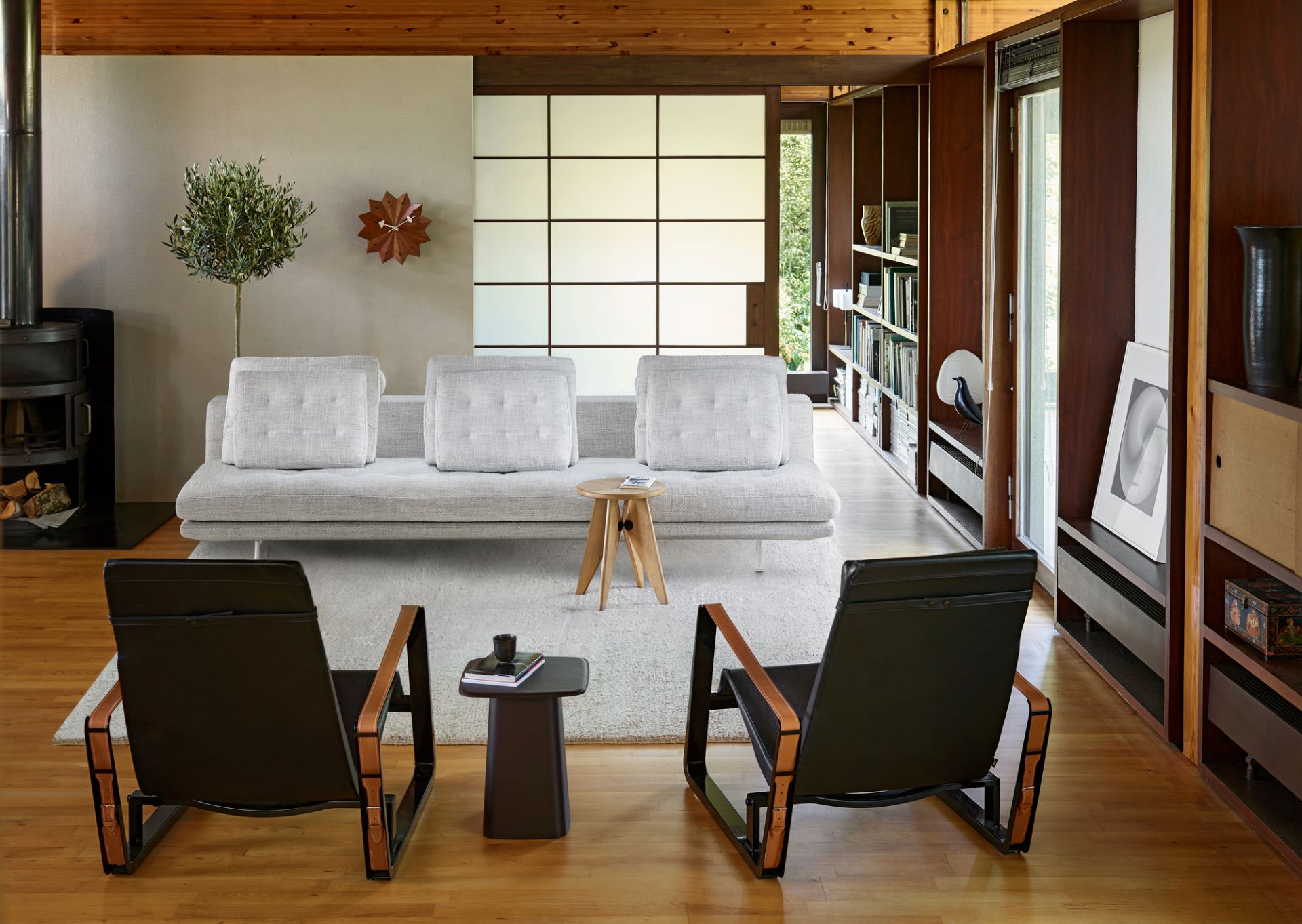 Modernes Wohnzimmer mit grauem Sofa, schwarzen Ledersesseln und Holztischen auf hellem Teppich.