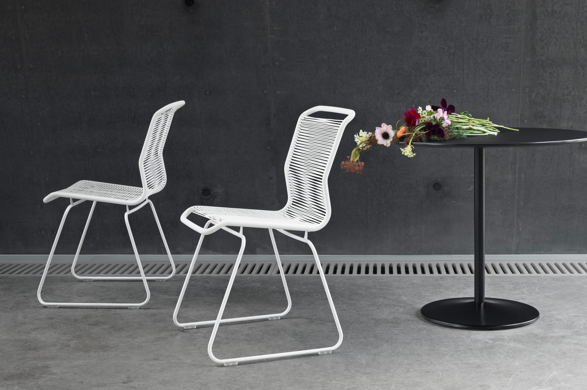 Zwei weiße Stühle und ein runder, schwarzer Montana Tisch mit Blumenstrauß vor grauer Wand.