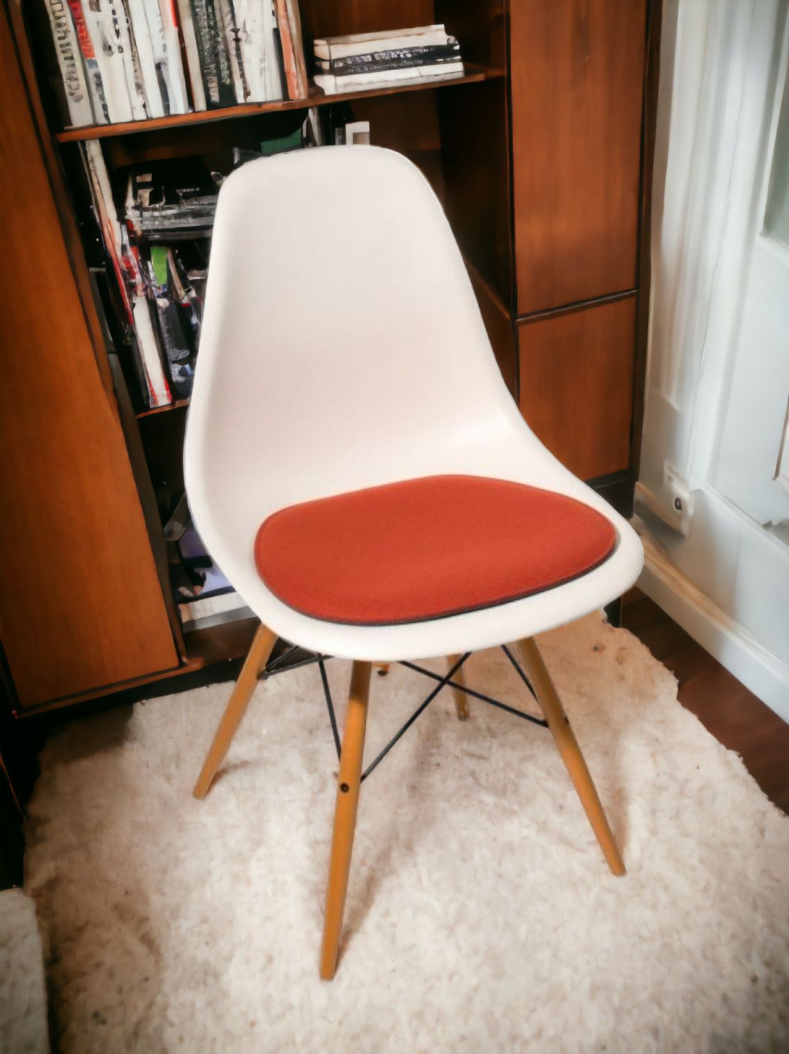 Weißer Design Stuhl mit Holzbeinen und rotem Sitzkissen vor Bücherregal und weißer Tür.