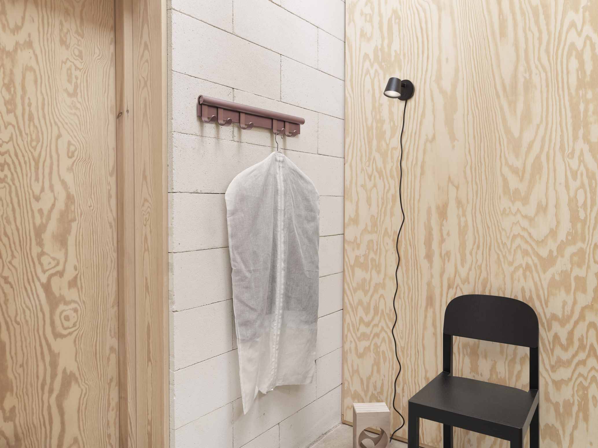 Muuto Coil Coat Rack Wandgarderobe aus braunem Metall mit Kleidersack an einer weißen Ziegelwand.