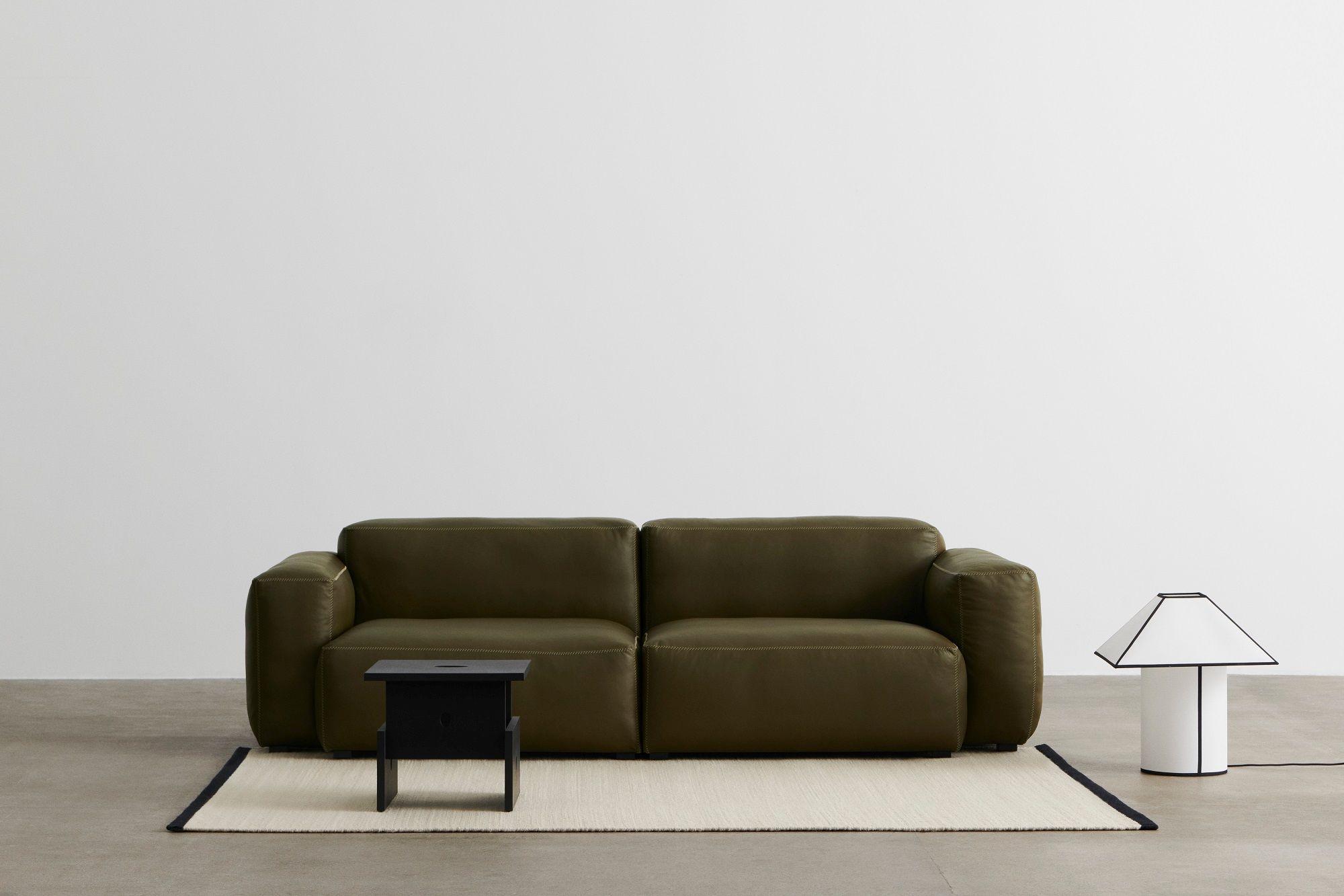 Ava Pyramid Tischleuchte von Hay, weiße Lampe mit schwarzem Rand neben olivgrünem Sofa.