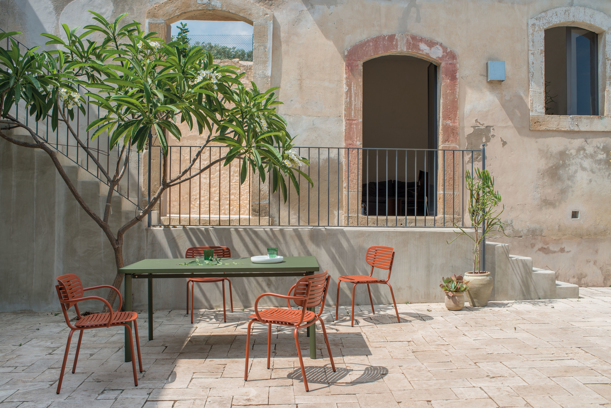 Mom Stuhl Emu: Gartenmöbel-Set mit orangefarbenen Stühlen und grünem Tisch auf sonnigem Innenhof.
