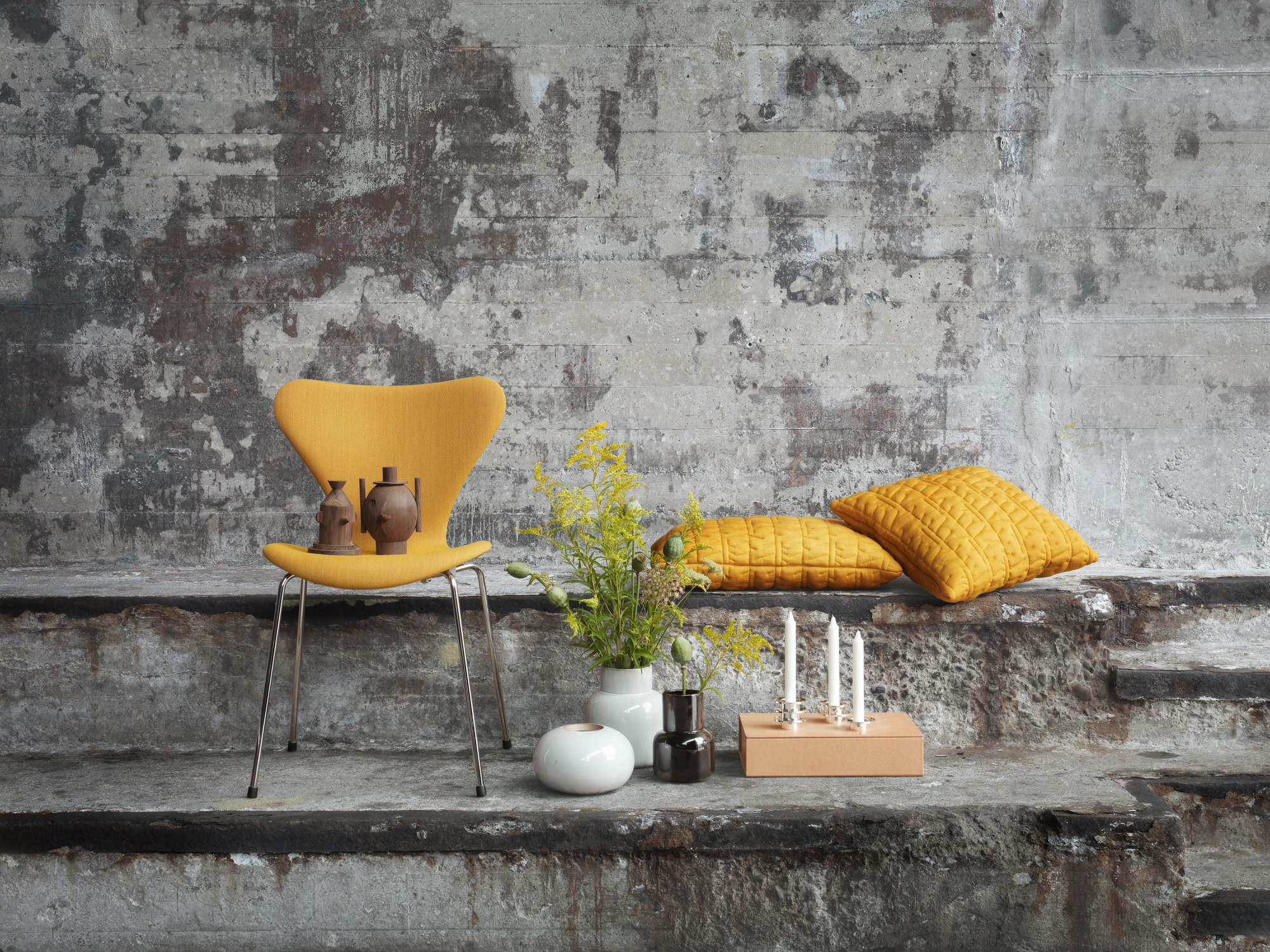 Dekoratives Stillleben mit Fritz Hansen Keramik Vase, gelbem Stuhl, Kissen und weiteren Wohnaccessoires vor Betonwand.