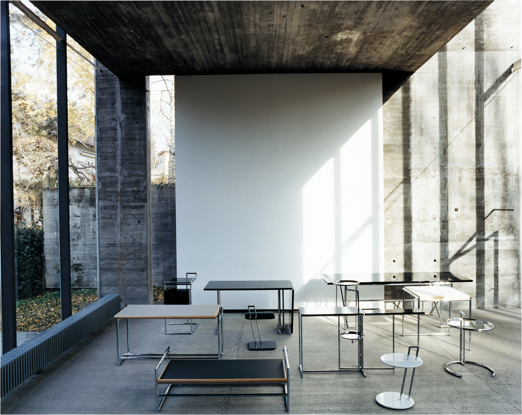 Verschiedene ClassiCon Tische, darunter der E1027 Beistelltisch, in einem modernen Raum mit Betonwänden. Designmöbel für Wohnen und Büro.