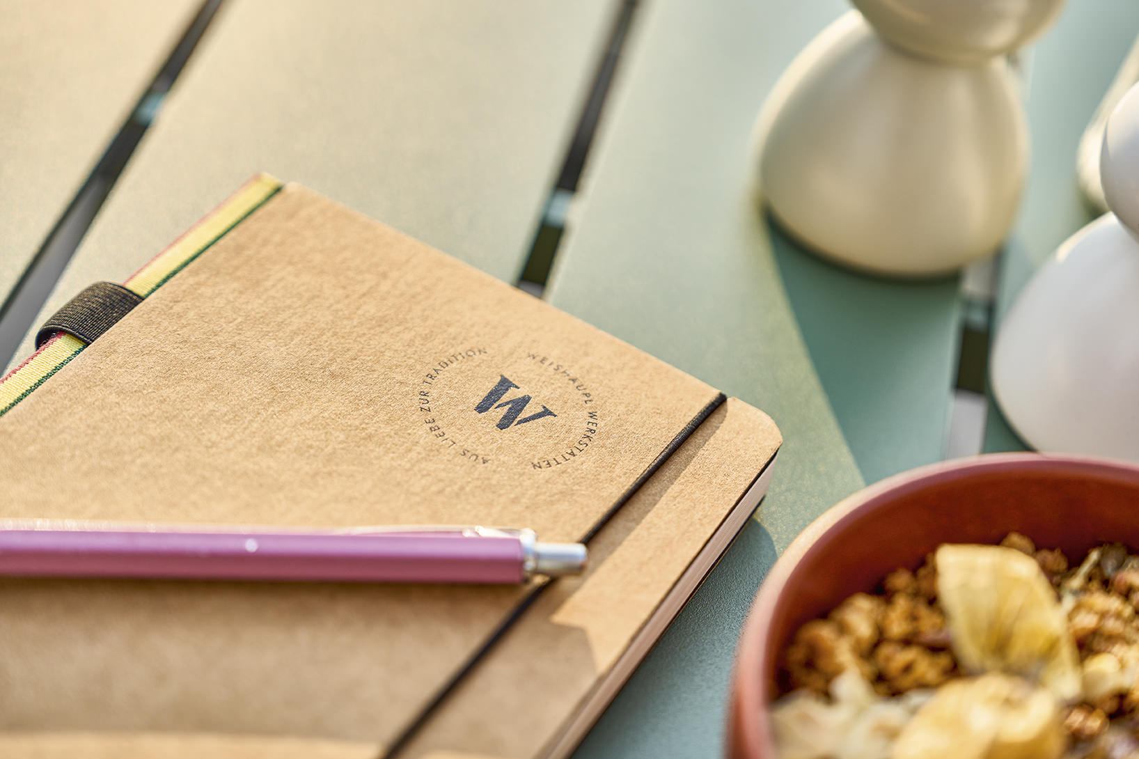 Detailaufnahme eines Weishäupl Vapio Outdoor Tisches mit Notizbuch, Stift, Schale mit Essen und Gewürzstreuer.
