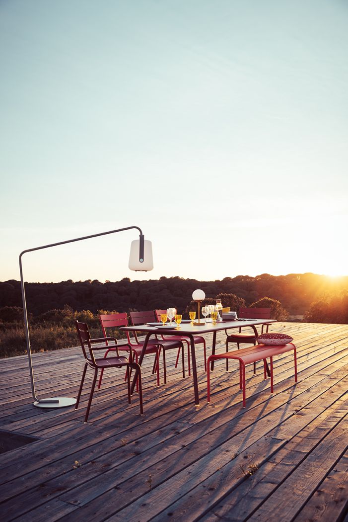 Roter Luxembourg Stuhl aus Stahl von Fermob auf einer Holzterrasse mit Esstisch und Beleuchtung.