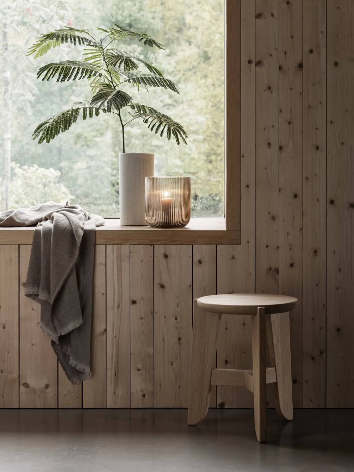 Blomus Eli Hocker aus hellem Holz vor Fenster mit Pflanze und Kerze.