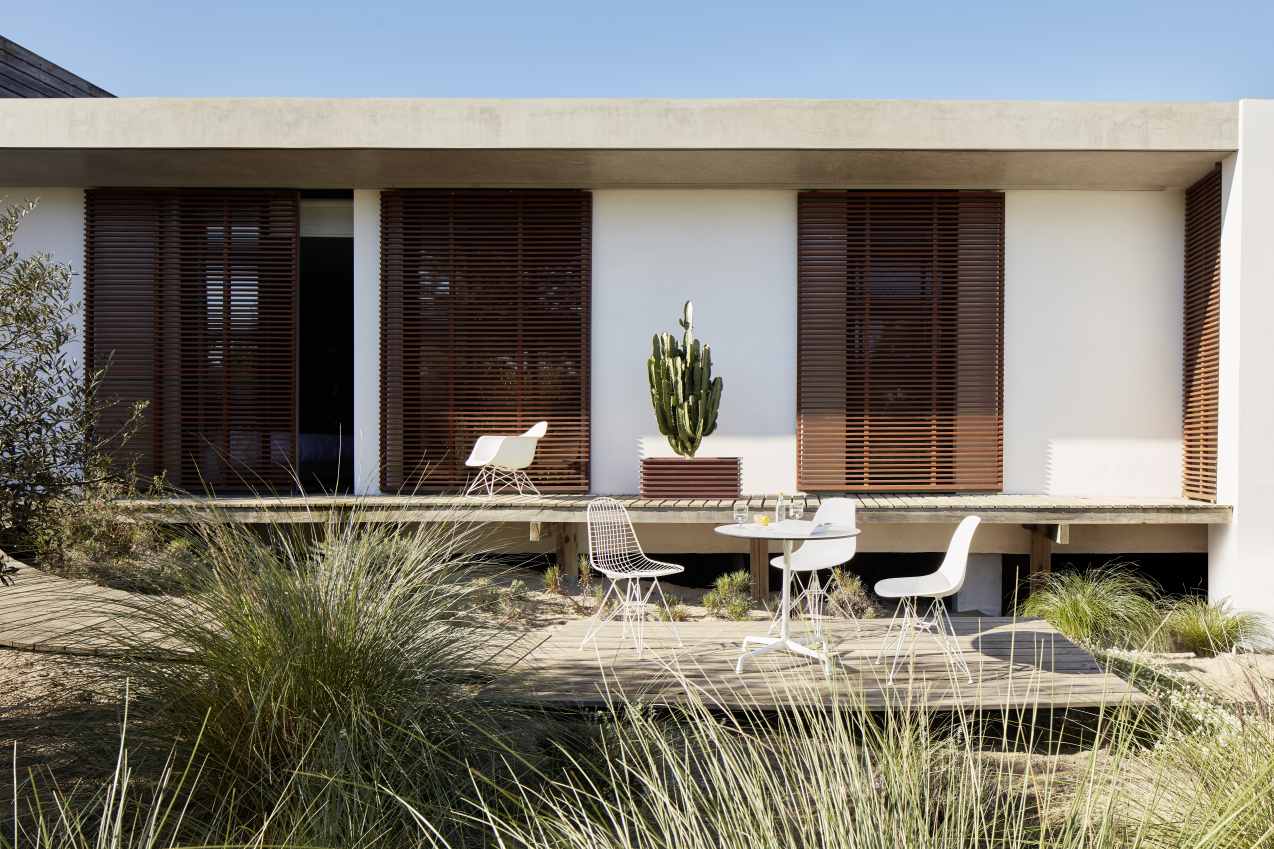 Vitra Eames Contract Table, runder Gartentisch mit weißen Stühlen auf einer Holzterrasse vor modernem Haus.