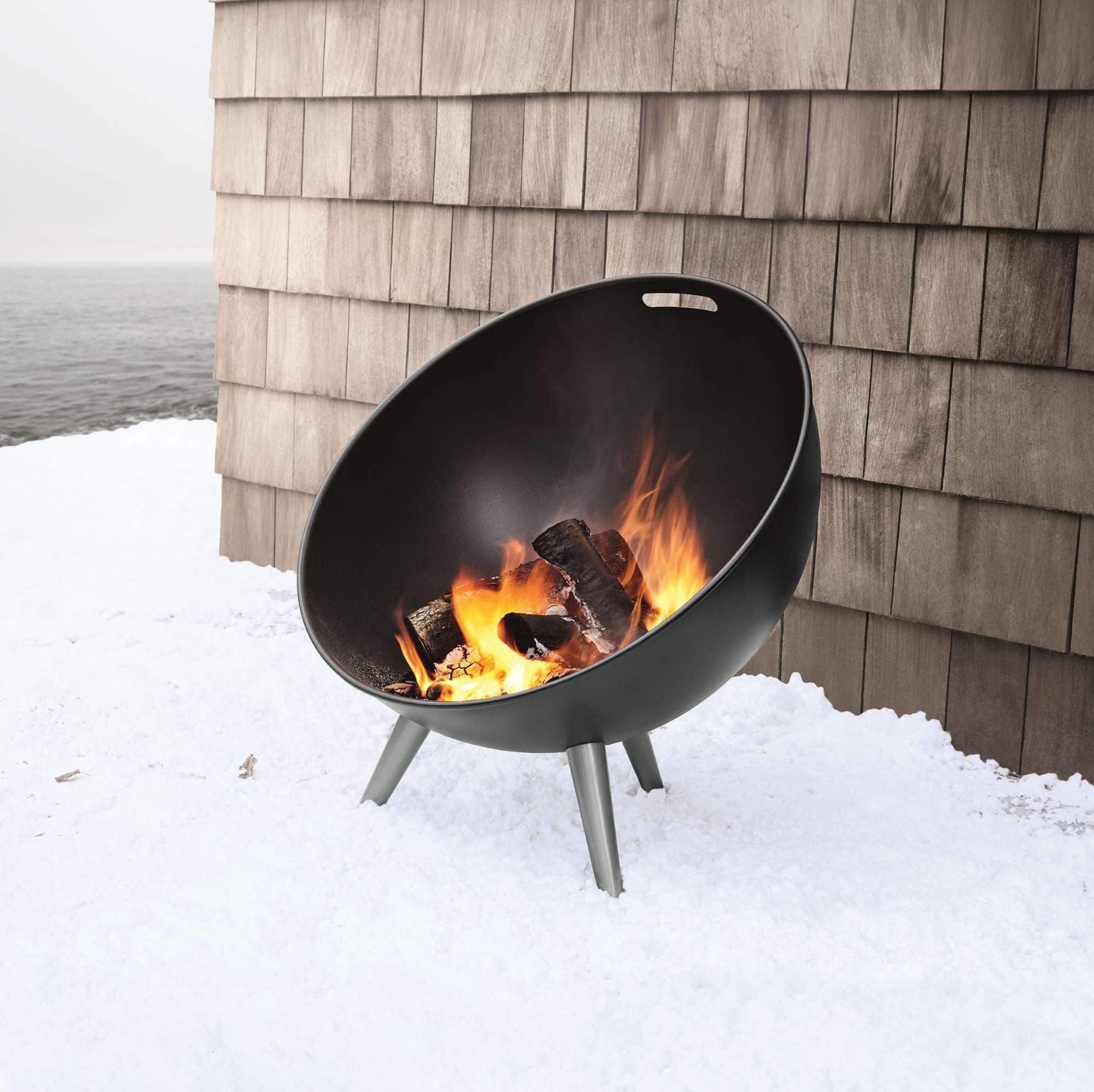 Eva Solo Fire Globe Feuerschale mit brennendem Holz auf Schnee vor einer Holzwand.