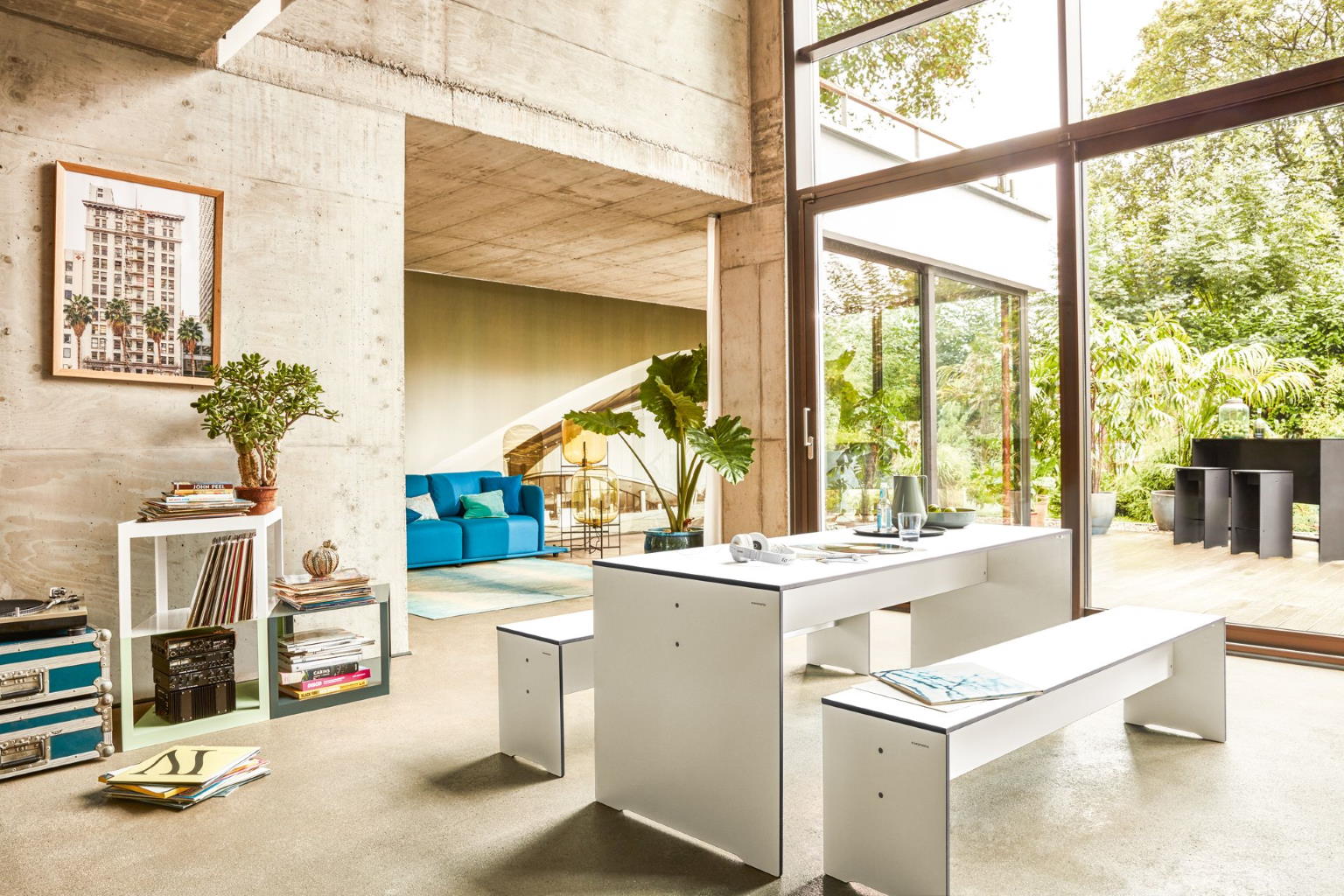 Heller Wohnraum mit weißem Esstisch und Bänken, blauer Couch und großen Fenstern zum Garten.