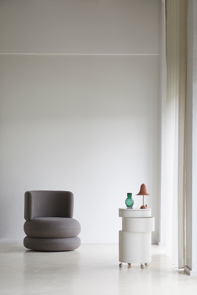Grauer Easy Chair Sessel mit runder Form, neben einem weißen Beistelltisch mit Lampe und Vase.