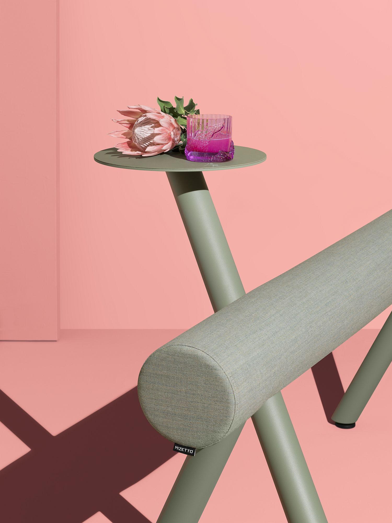 Mizetto Lumber Floor Sitzmöbel in Grün mit Beistelltisch, Protea-Blüte und Getränk vor rosa Hintergrund.