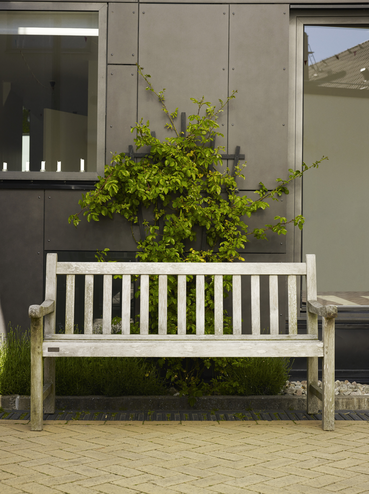 Helle Holzbank "England Bench" von Skagerak im Außenbereich vor Hauswand mit Grünpflanzen.