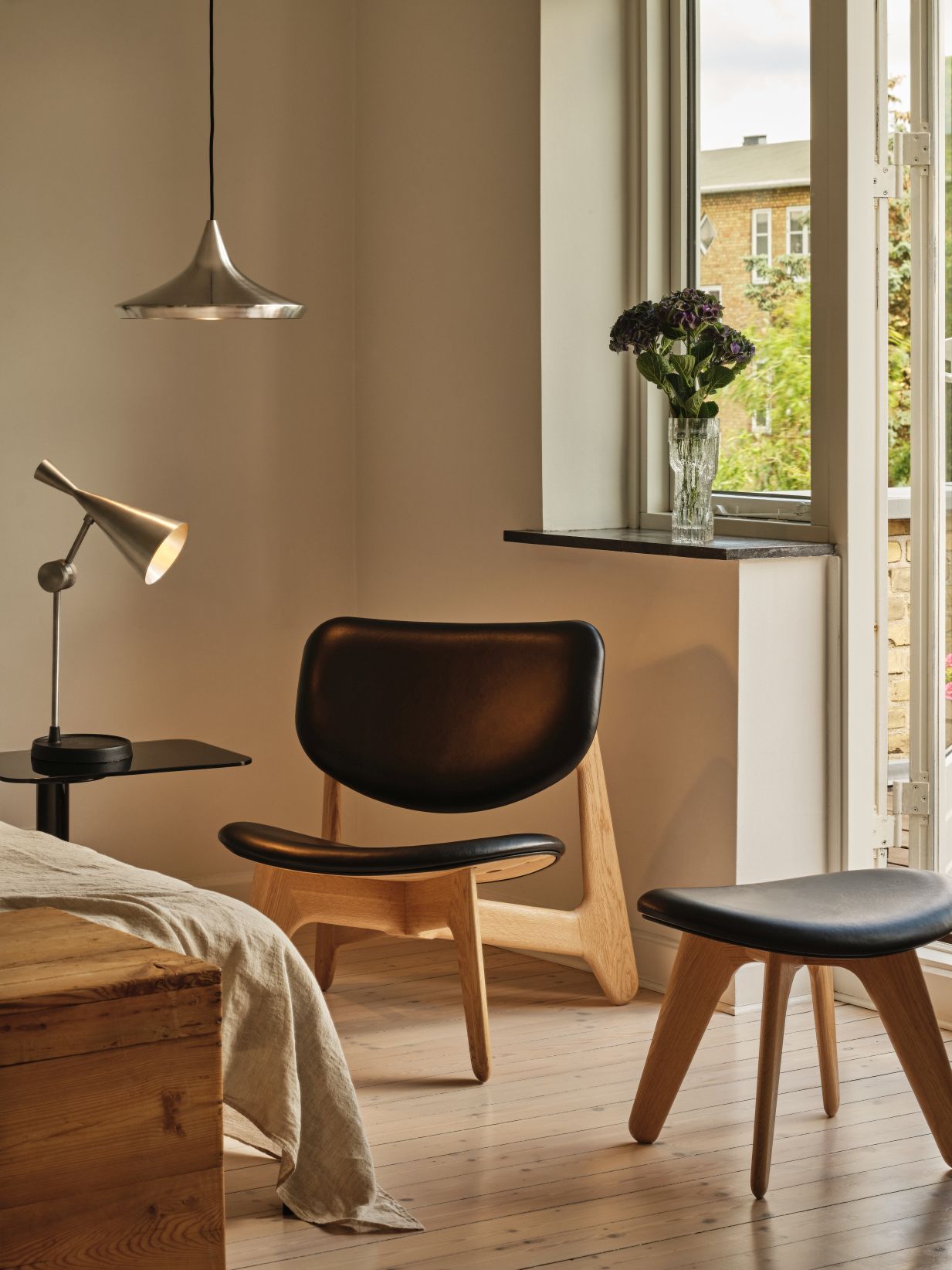 Helle Raumansicht mit Tom Dixon Unbeaten Wide Pendelleuchte, Tischleuchte, Holzstuhl, Hocker und Fenster mit Blumen.