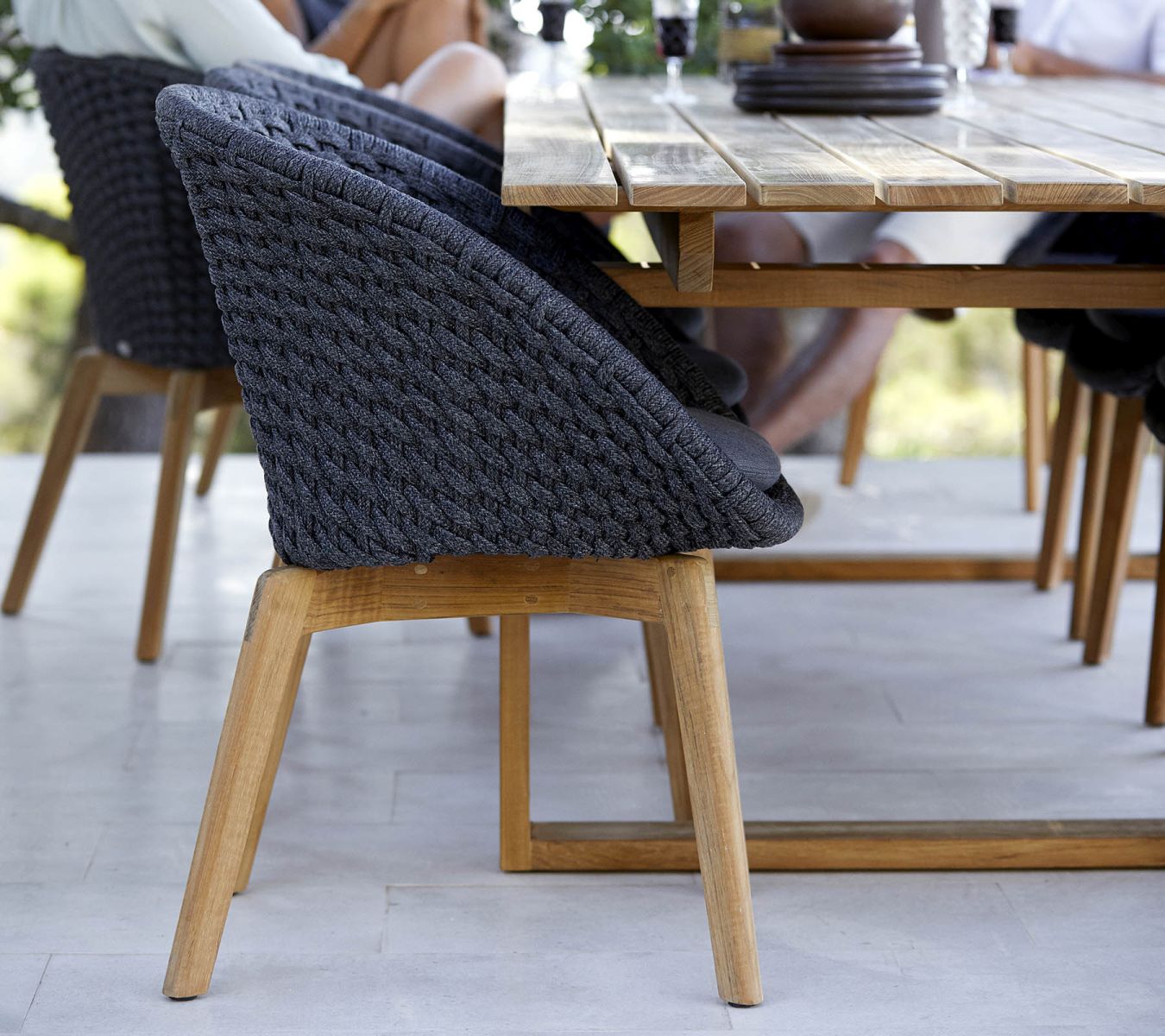 Detailaufnahme des Cane-Line Endless Esstisches aus Teakholz mit grauen Flechtstühlen im Außenbereich.