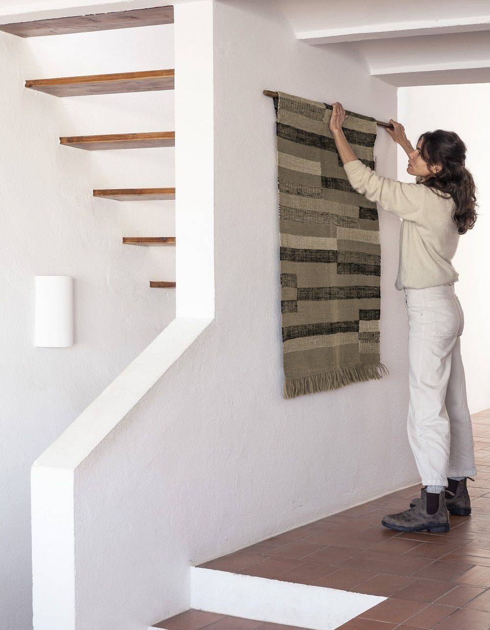 Frau hängt gewebten Wandteppich mit geometrischem Muster an einer Holzstange an einer weißen Wand.